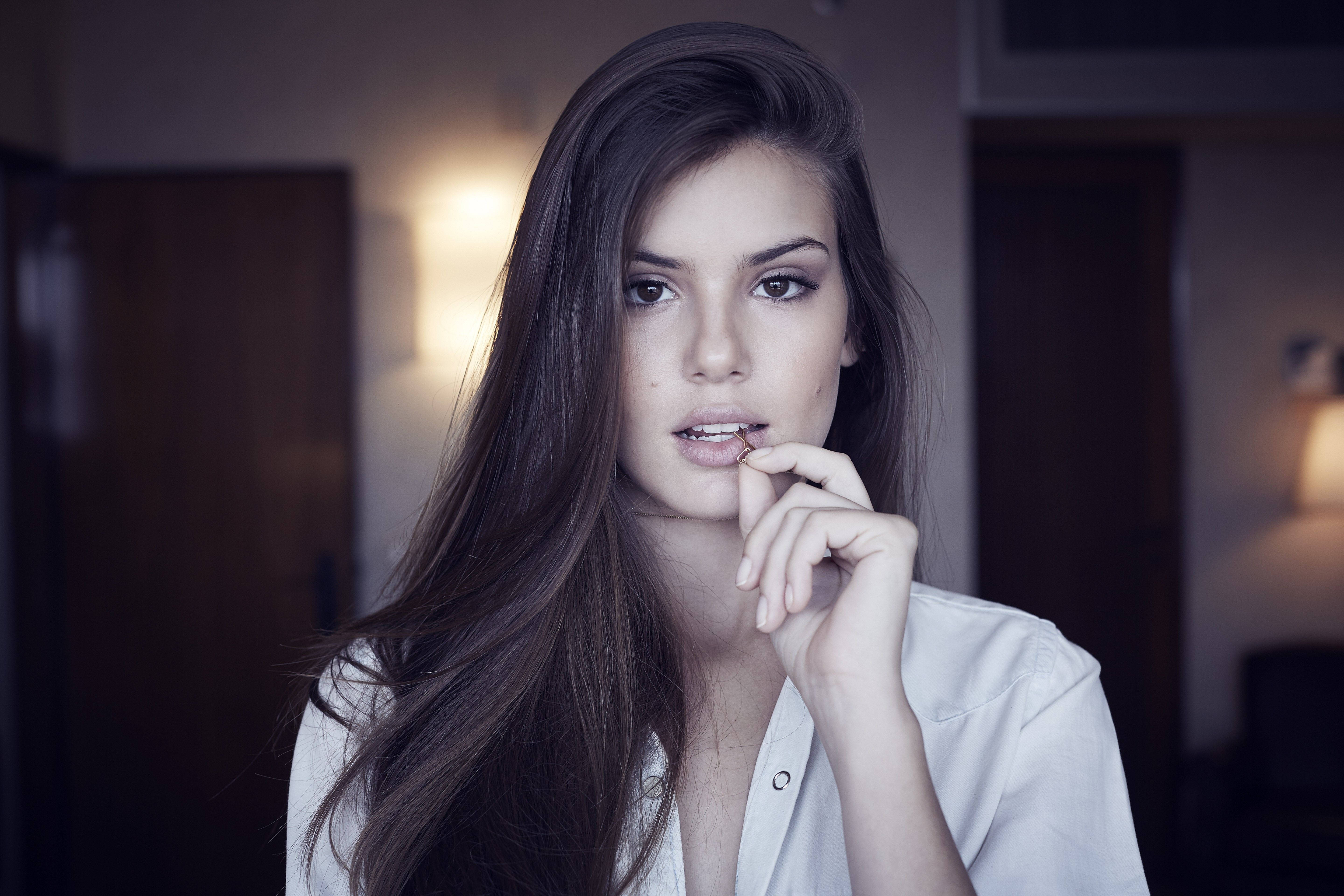 Woman Beautiful 桌布 壁紙 — 一位長黑髮的年輕女性在柔和光線的室內環境中，將手指放在嘴唇附近，若有所思地看著鏡頭。