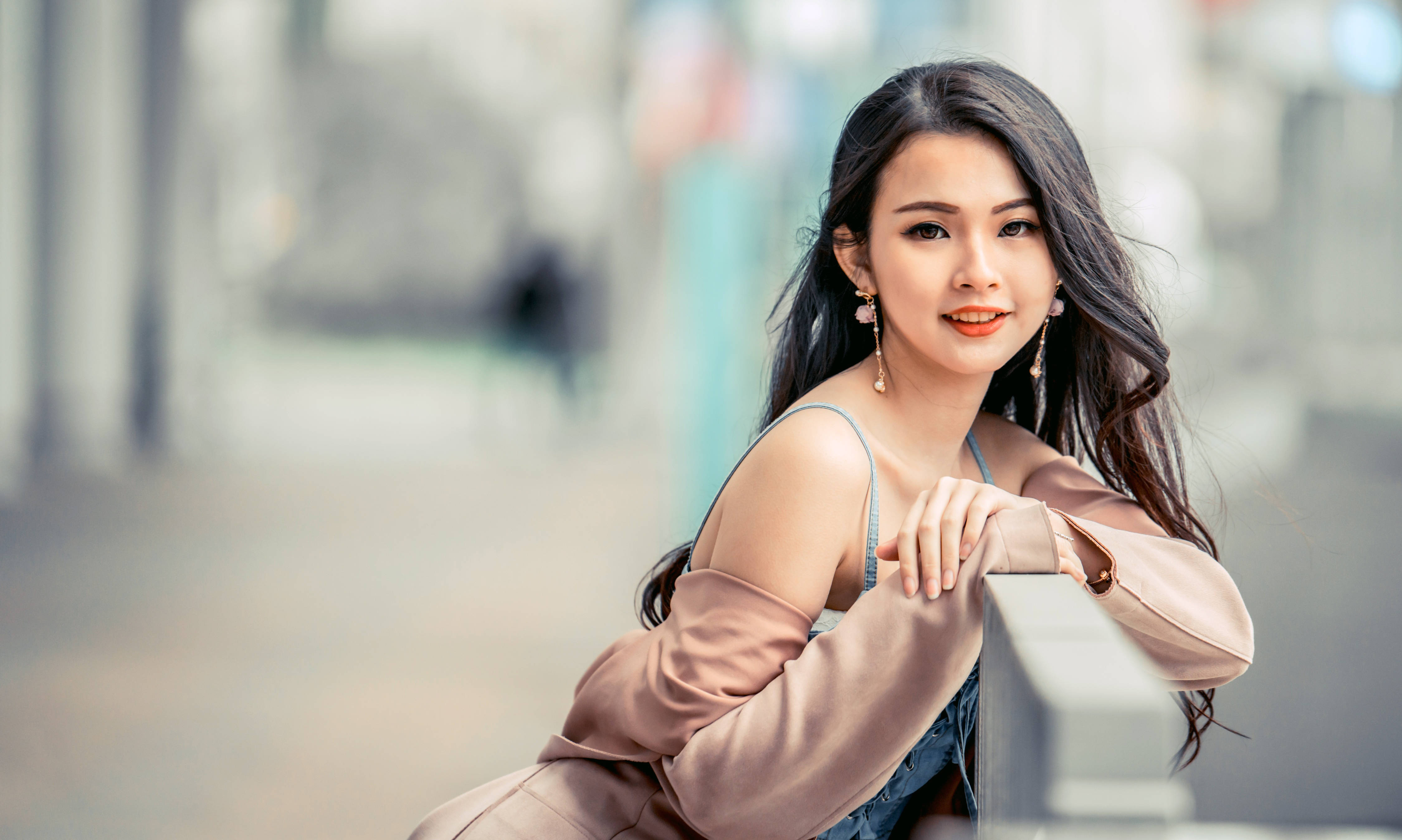 Woman 桌布 壁紙 — 一位長黑髮的年輕女性在柔和模糊的城市街道上倚靠欄杆，露出溫暖的笑容。