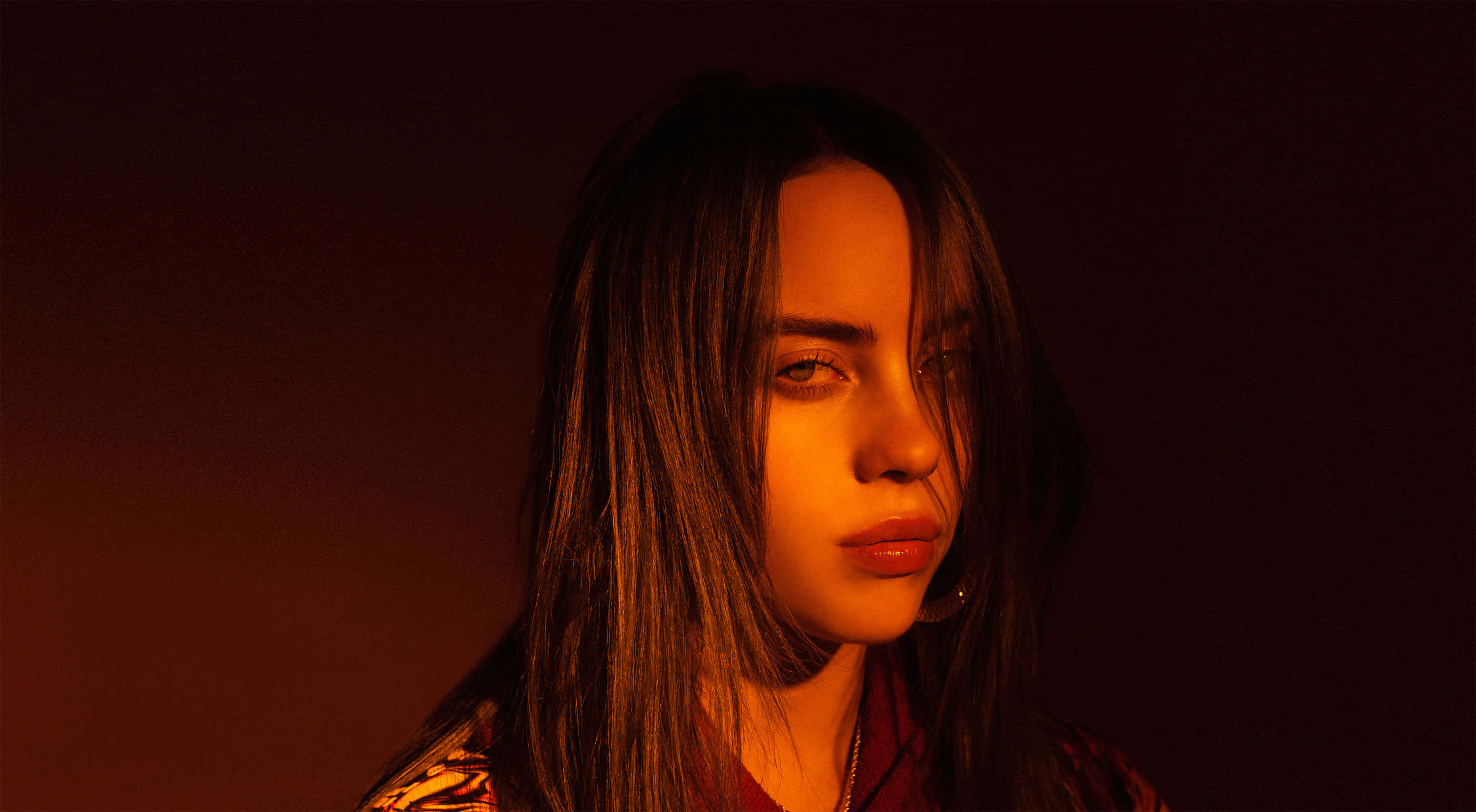 Billie Eilish Laptop 桌布 壁紙 — 一位長黑髮的年輕女性在溫暖的橙色燈光下，對著深紅色背景柔和地凝視。