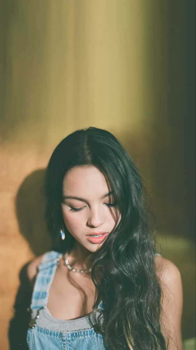 Olivia Rodrigo Aesthetic 桌布 壁紙 — 一位長黑髮的年輕女性在溫暖的金色燈光下穿著牛仔吊帶褲和珍珠項鍊。