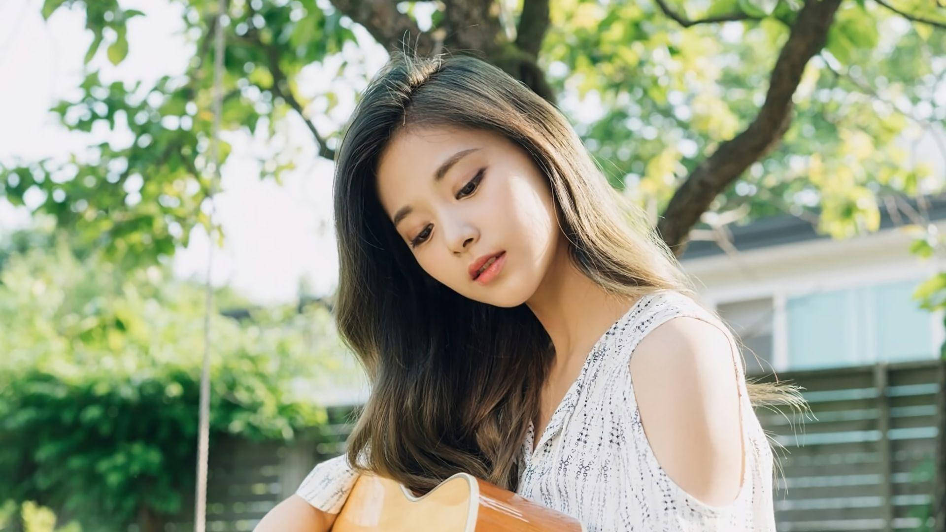 子瑜 桌布 壁紙 — 一位長黑髮的年輕女性在茂密綠樹間灑落的斑駁陽光下，低頭抱著一把木吉他。