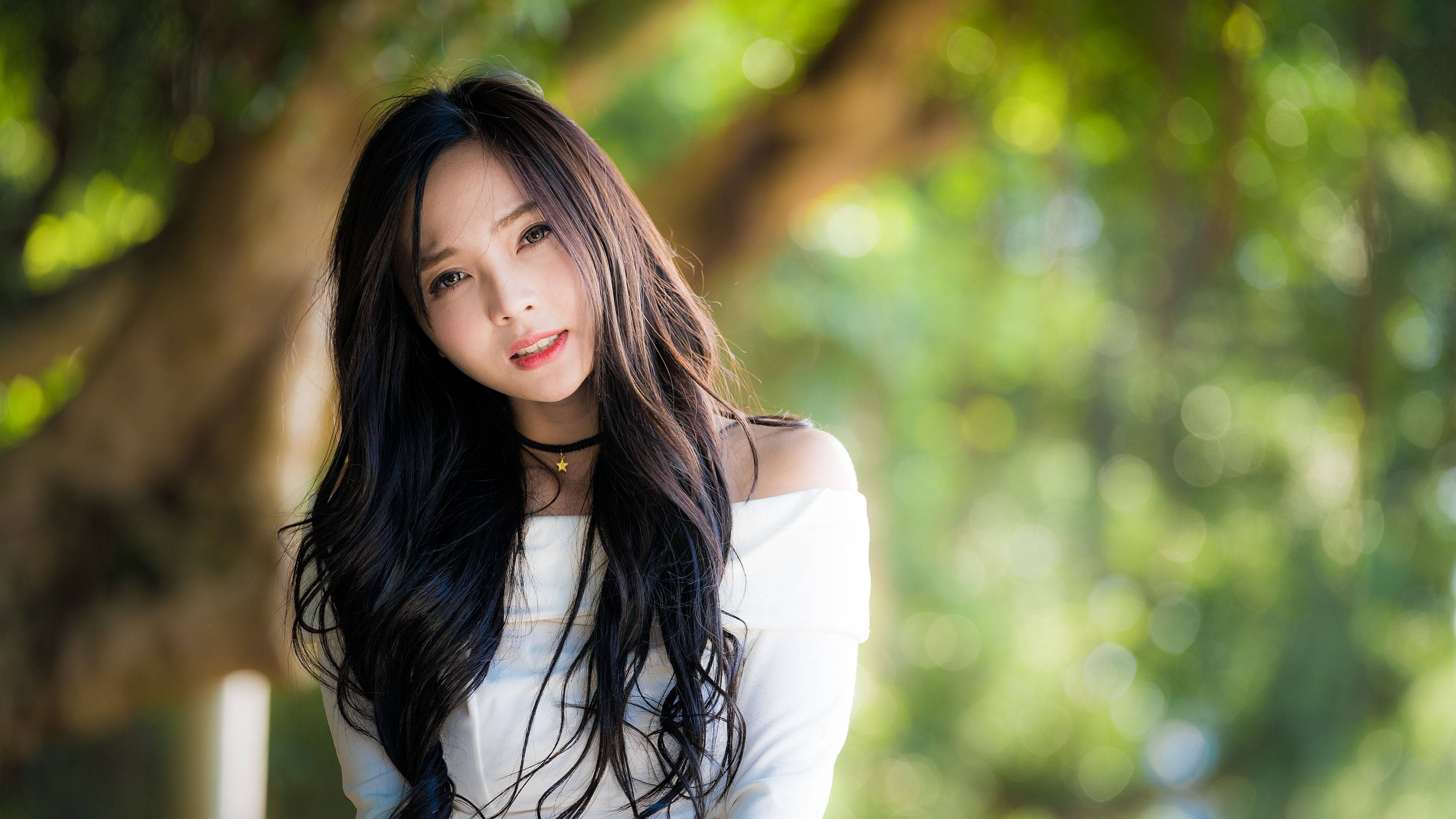 美女 桌布 壁紙 — 一位長黑髮的年輕女性在陽光照射的樹下微笑，背景是模糊的綠色。