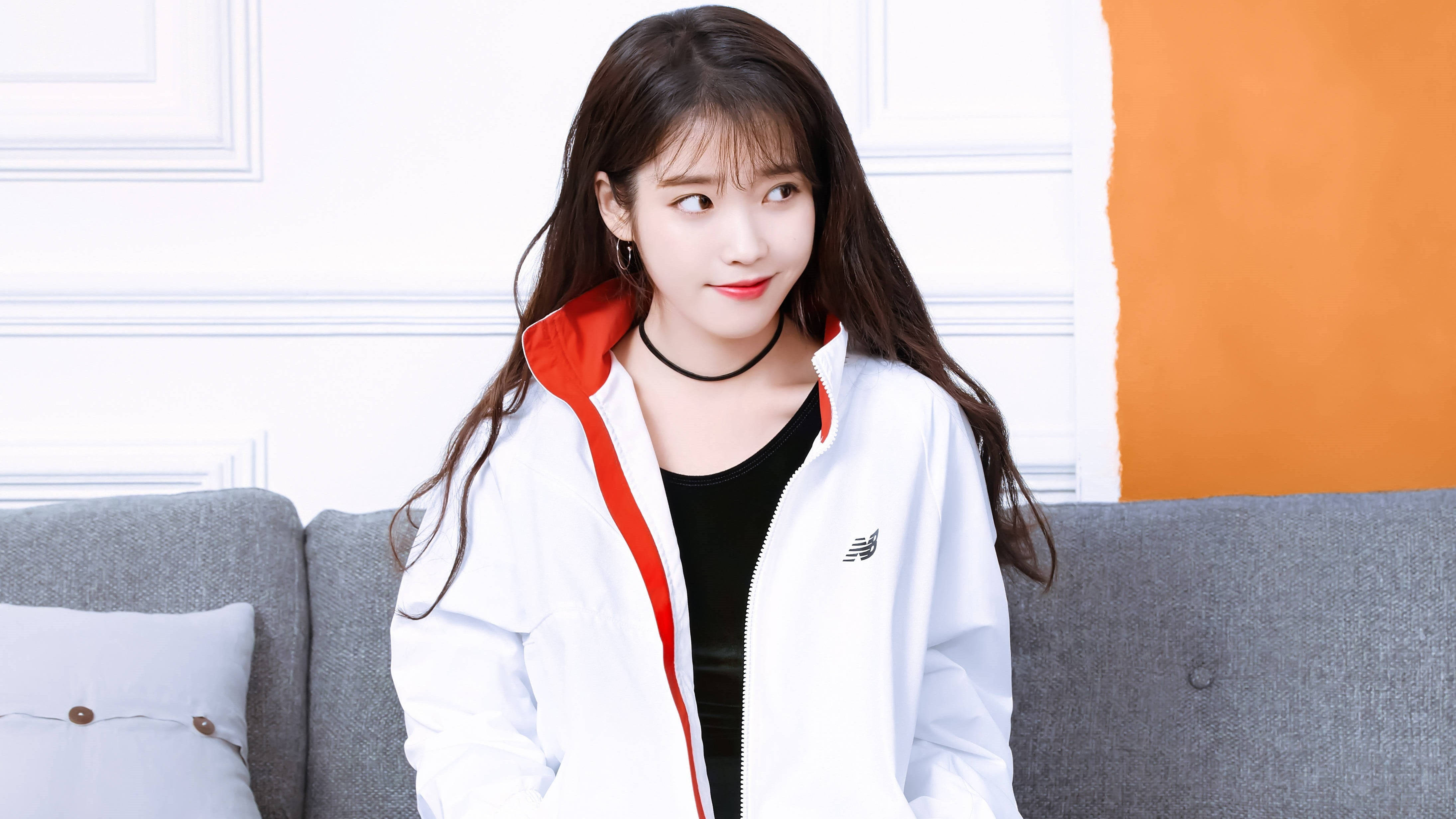 Kpop Aesthetic 桌布 壁紙 — 一位長黑髮的年輕女性坐在灰色沙發上，身穿帶紅色飾邊的白色外套和黑色上衣。