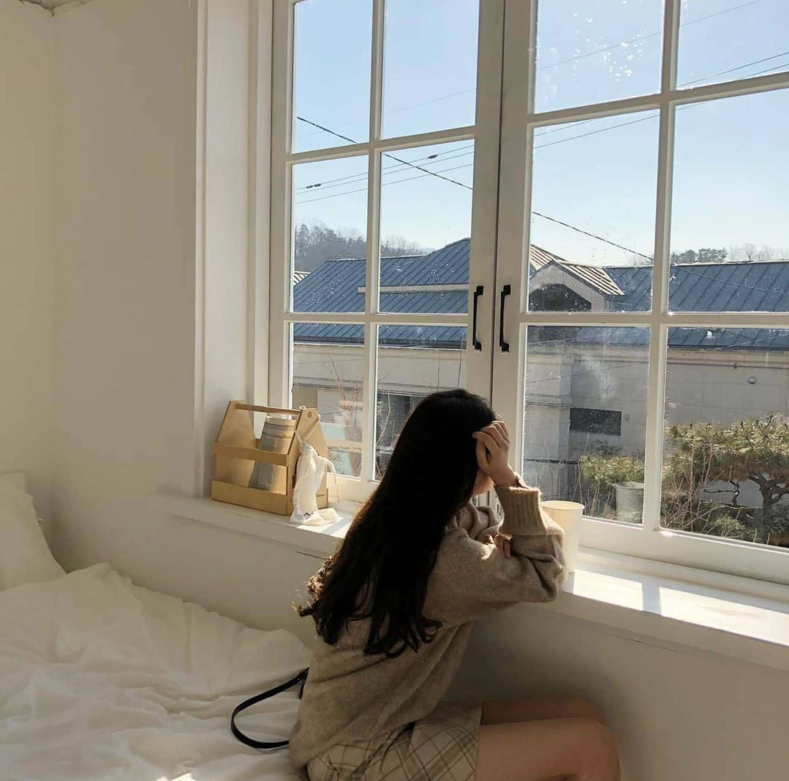 美學米色 桌布 壁紙 — 一位長黑髮的年輕女性坐在簡約臥室的明亮窗邊，凝視著晴朗藍天下屋頂的景色。