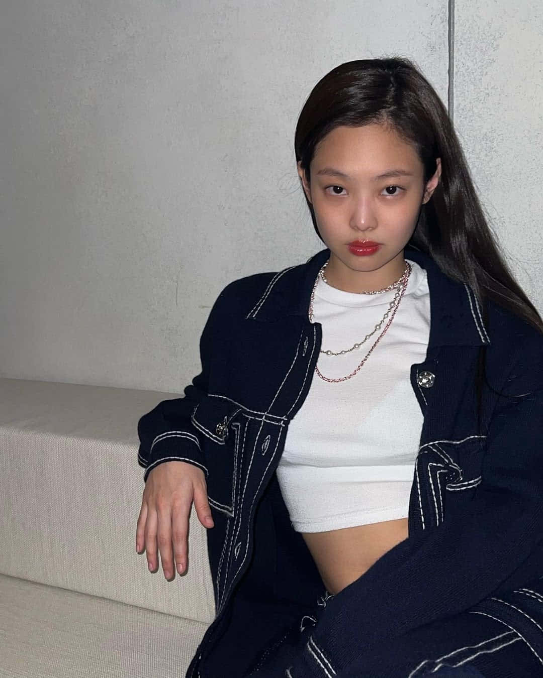BLACKPINK 桌布 壁紙 — 一位長黑髮的年輕女性坐在米色沙發上，身穿白色車縫線的深藍色外套，內搭白色短版上衣，佩戴多層項鍊。