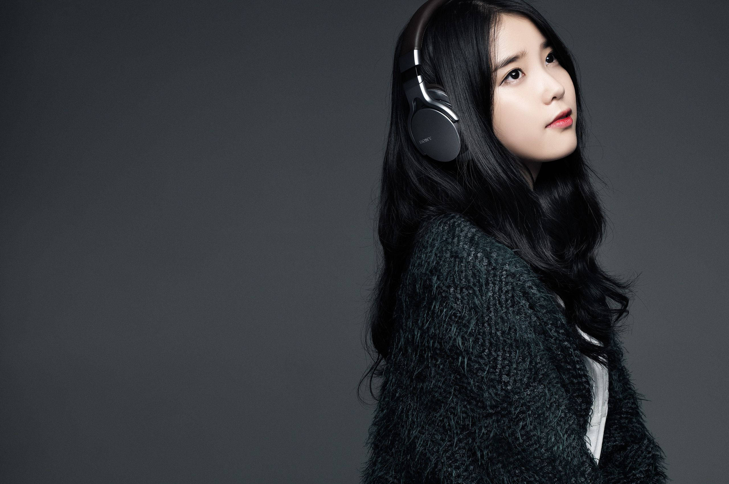 Kpop Aesthetic 桌布 壁紙 — 一位長黑髮的年輕女性戴著頭戴式耳機和質感深色毛衣，背景是純灰色攝影棚。