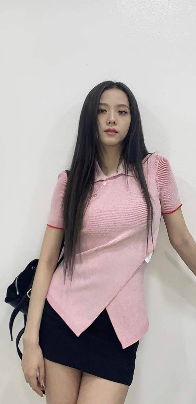 Jisoo Cute 桌布 壁紙 — 一位長黑髮的年輕女性穿著帶紅色飾邊的粉紅色質感Polo衫和黑色迷你裙，靠著純白牆壁。