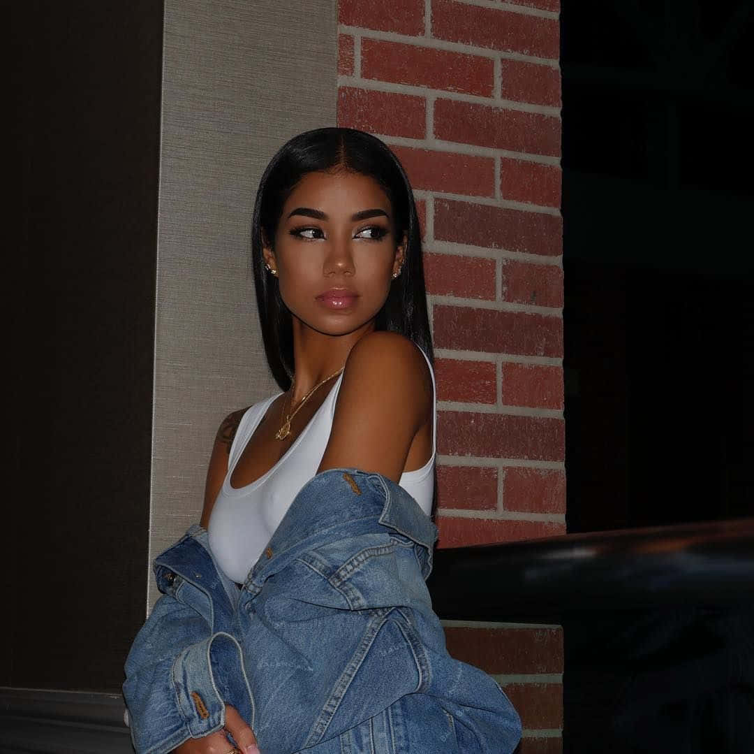 jhene aiko 桌布 壁紙 — 一位長黑髮的年輕女性穿著白色背心和牛仔外套靠在磚牆上。