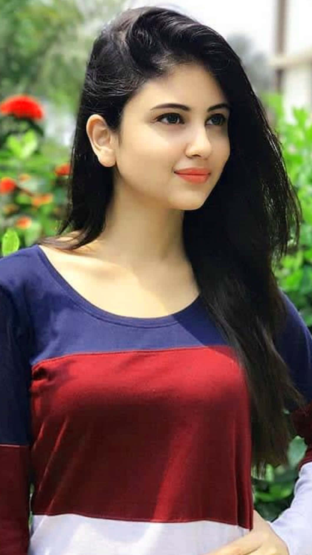 Beautiful Girl 桌布 壁紙 — 一位長黑髮的年輕女性穿著紅藍白條紋上衣，在茂密綠色花園背景下溫柔微笑。
