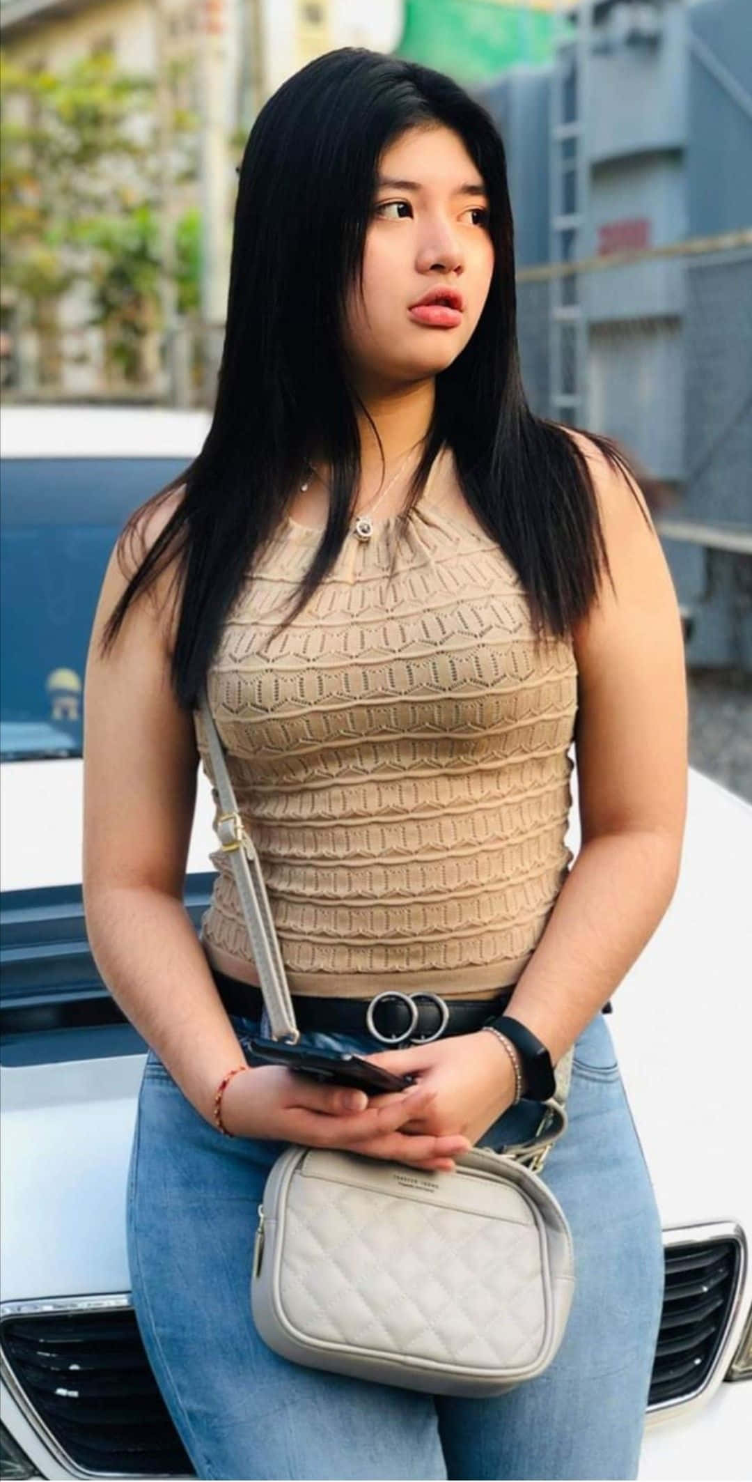 Jeans 桌布 壁紙 — 一位長黑髮的年輕女性站在白色汽車旁，身穿米色質感上衣和藍色牛仔褲，手持手機和絎縫斜背包。