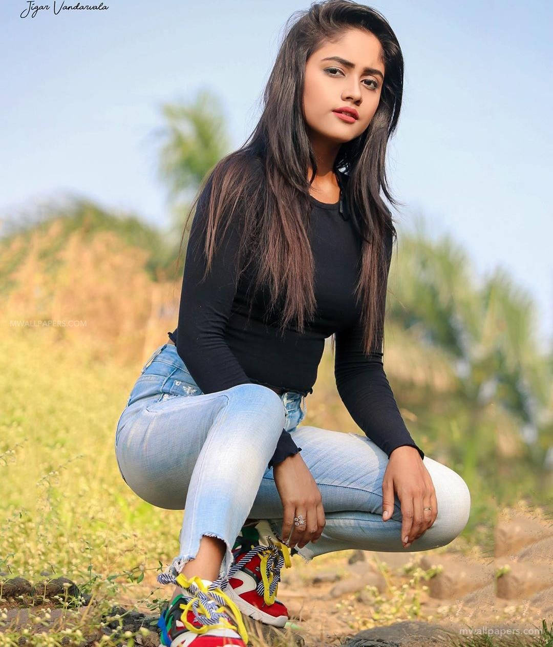 Nisha 桌布 壁紙 — 一位長黑髮的年輕女性蹲在陽光明媚的田野中，身穿黑色上衣、淺藍色牛仔褲和彩色運動鞋。
