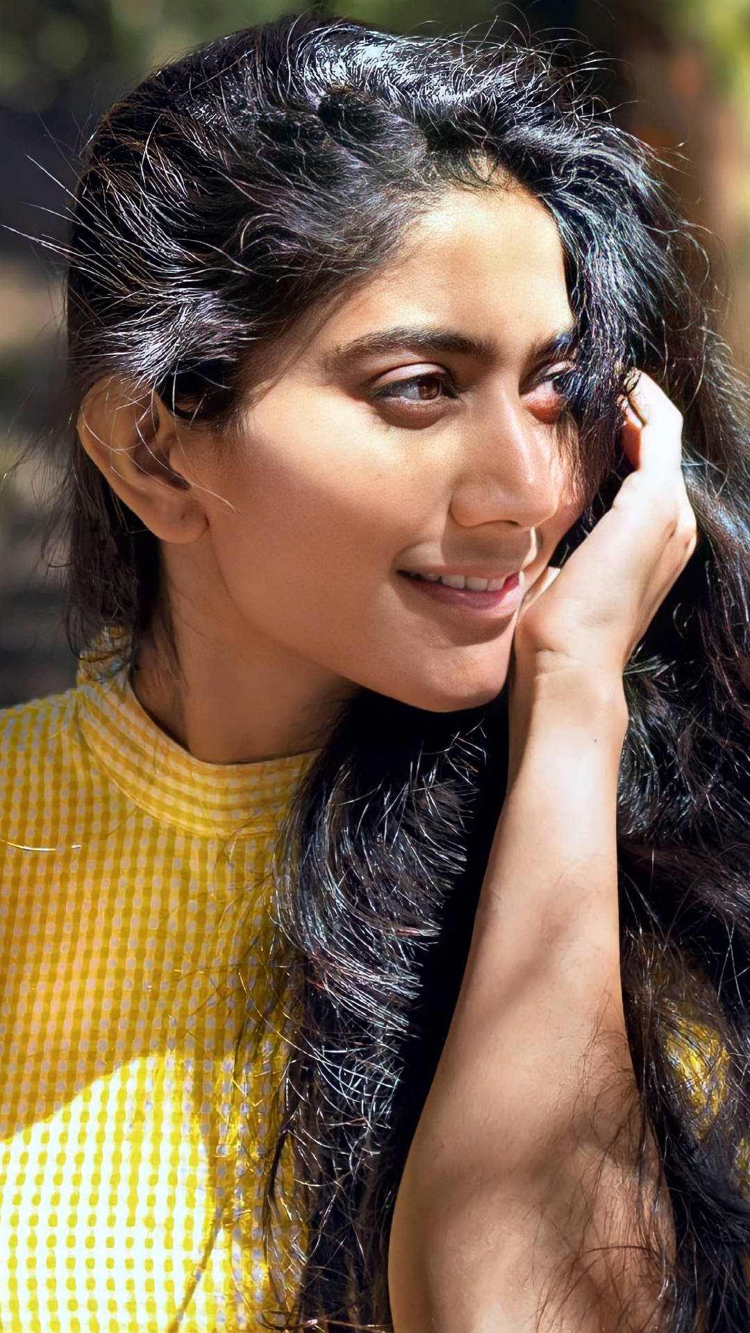 Sai Pallavi 桌布 壁紙 — 一位長黑髮的年輕女性輕觸臉龐，溫柔微笑，在明亮陽光下穿著黃色格紋上衣。