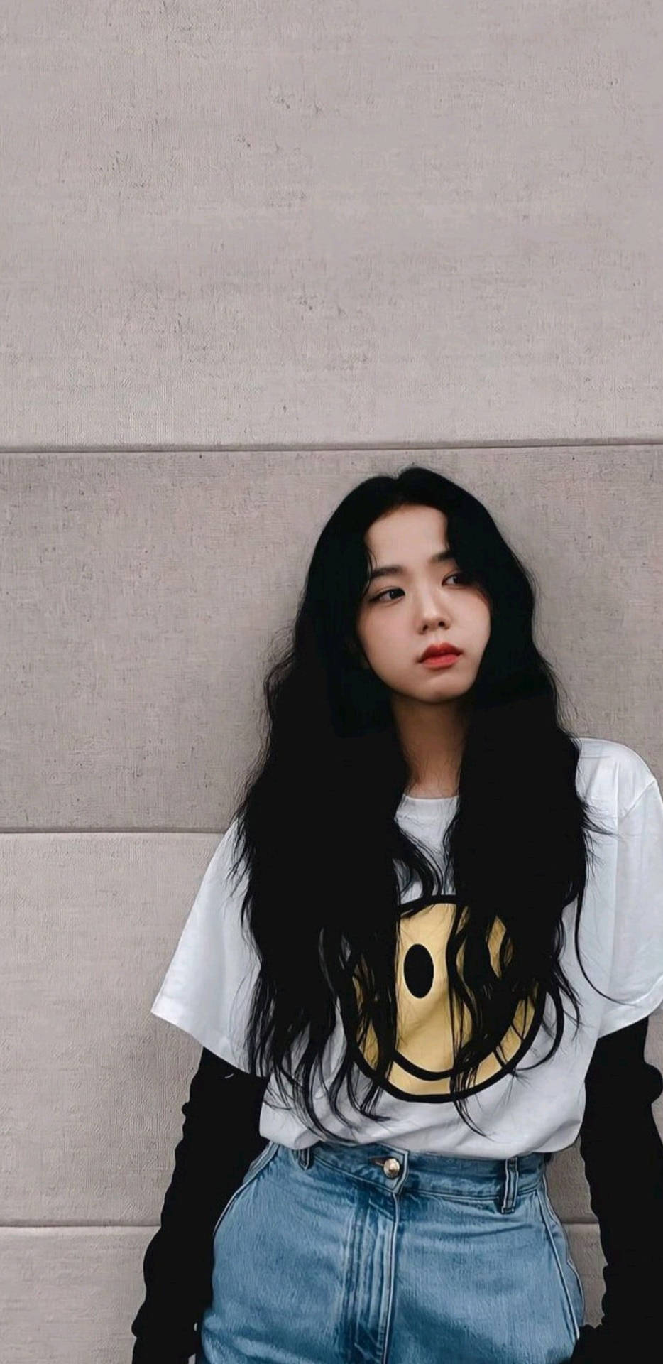 長黑髮的年輕女性穿著笑臉T恤和牛仔褲靠著混凝土牆。 — 智秀 GIF 壁紙