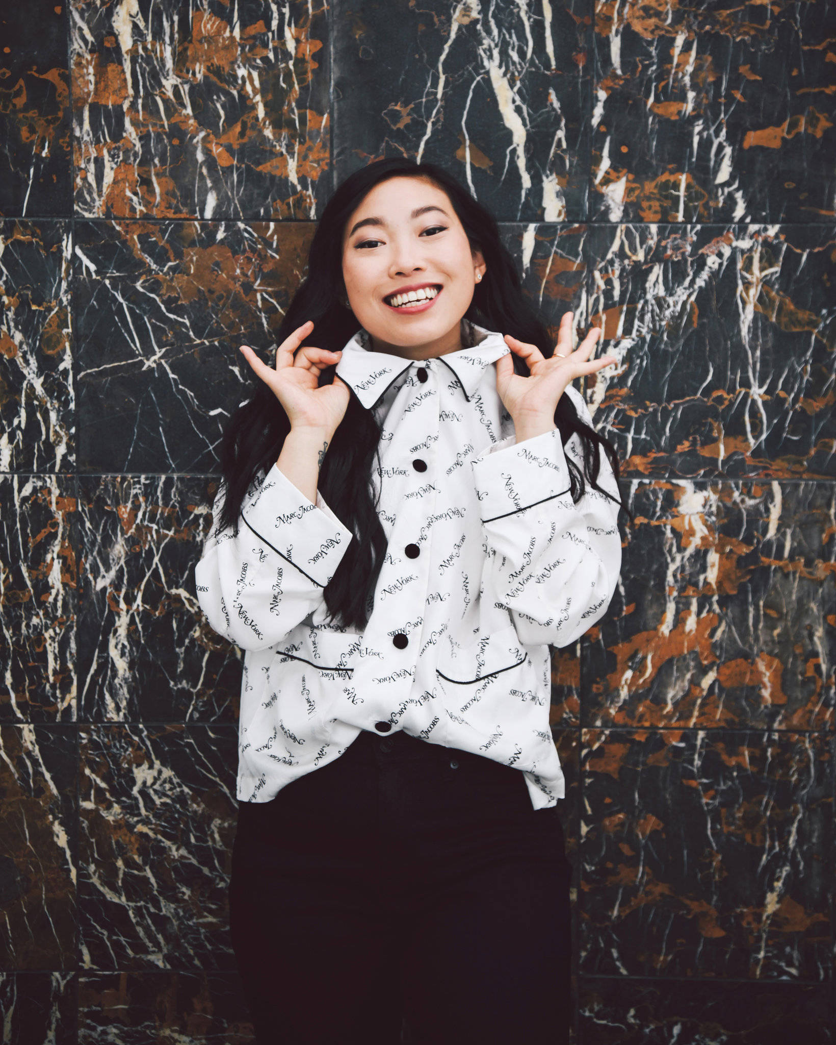 Awkwafina 桌布 壁紙 — 一位長黑髮的快樂女性，在引人注目的黑白金大理石牆前燦爛微笑擺姿勢。