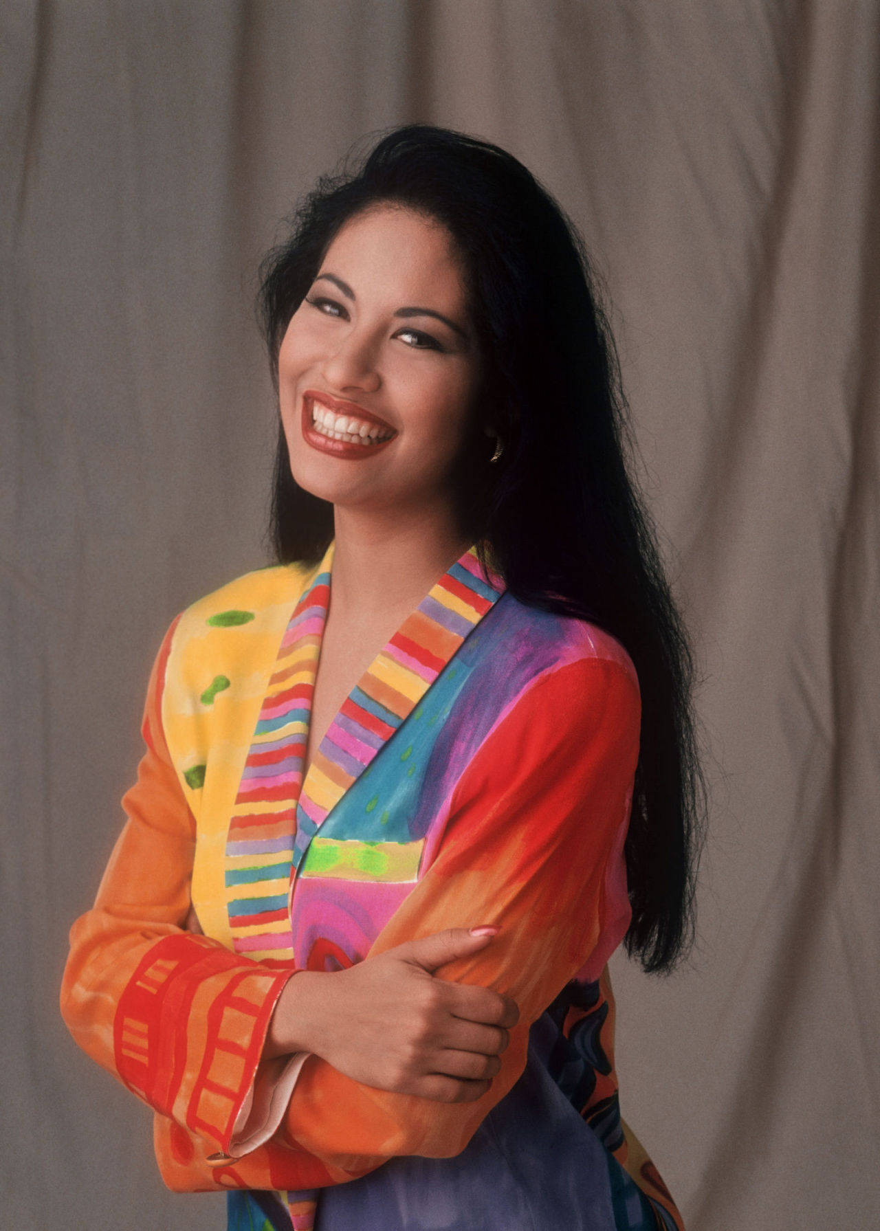 Selena Quintanilla 桌布 壁紙 — 一位長黑髮的燦爛女性，在中性工作室背景下穿著鮮豔多彩圖案外套，露出燦爛笑容。