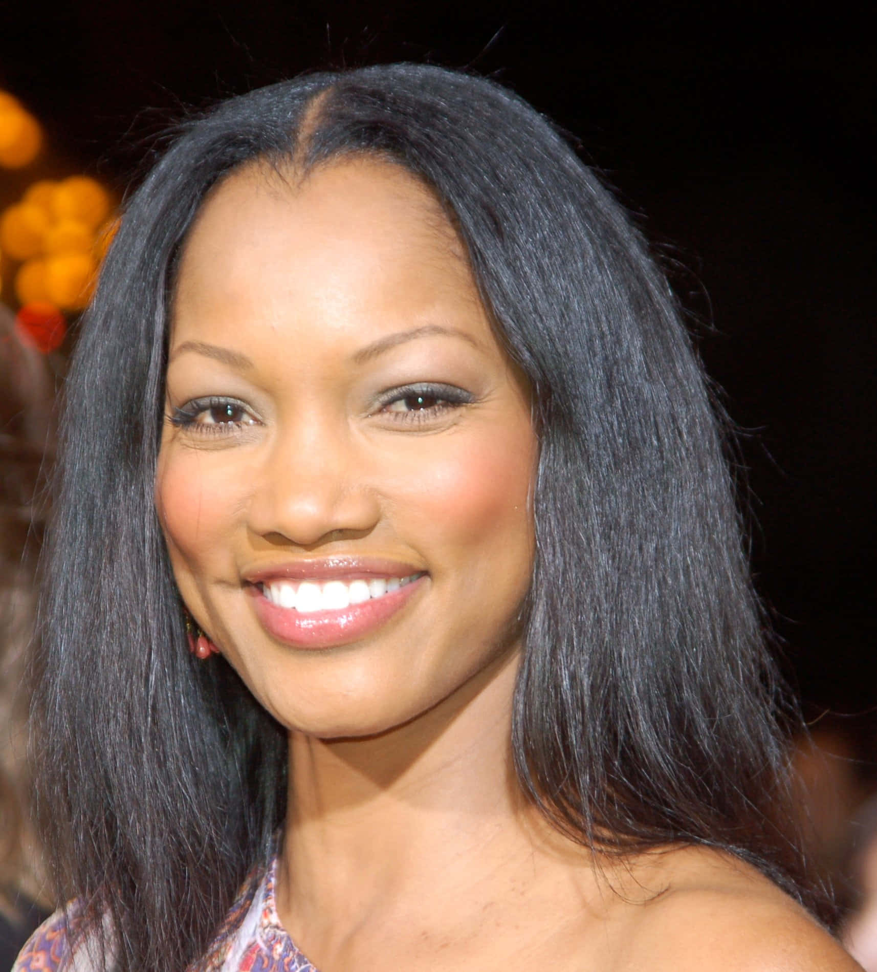 Garcelle Beauvais 桌布 壁紙 — 一位長黑髮的燦爛女性，在柔和模糊的金色燈光背景下溫暖微笑。