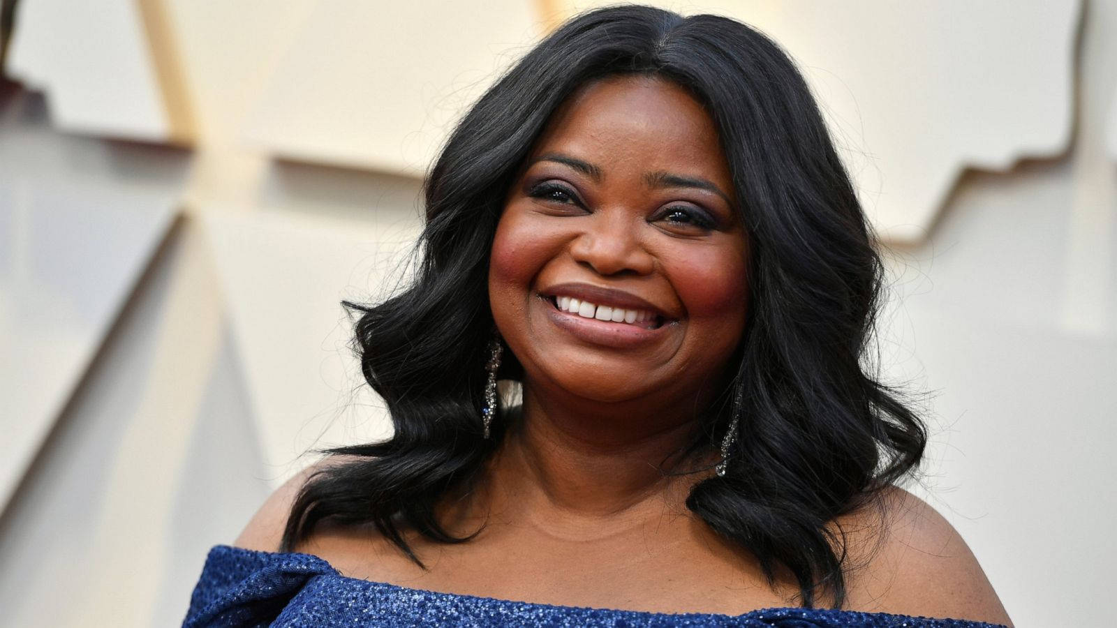 oscars 桌布 壁紙 — 一位長黑髮的燦爛女性，在華麗活動中穿著閃亮藍色露肩連衣裙，露出燦爛笑容。