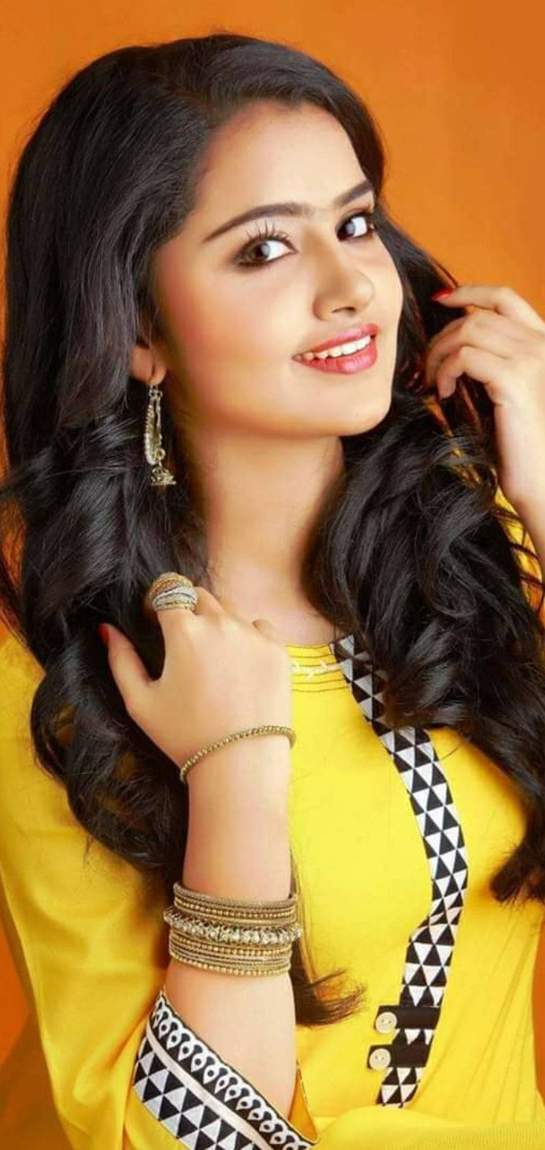 anupama parameswaran 桌布 壁紙 — 一位長黑髮的燦爛女性在溫暖的橙色背景前微笑著觸摸她的頭髮。
