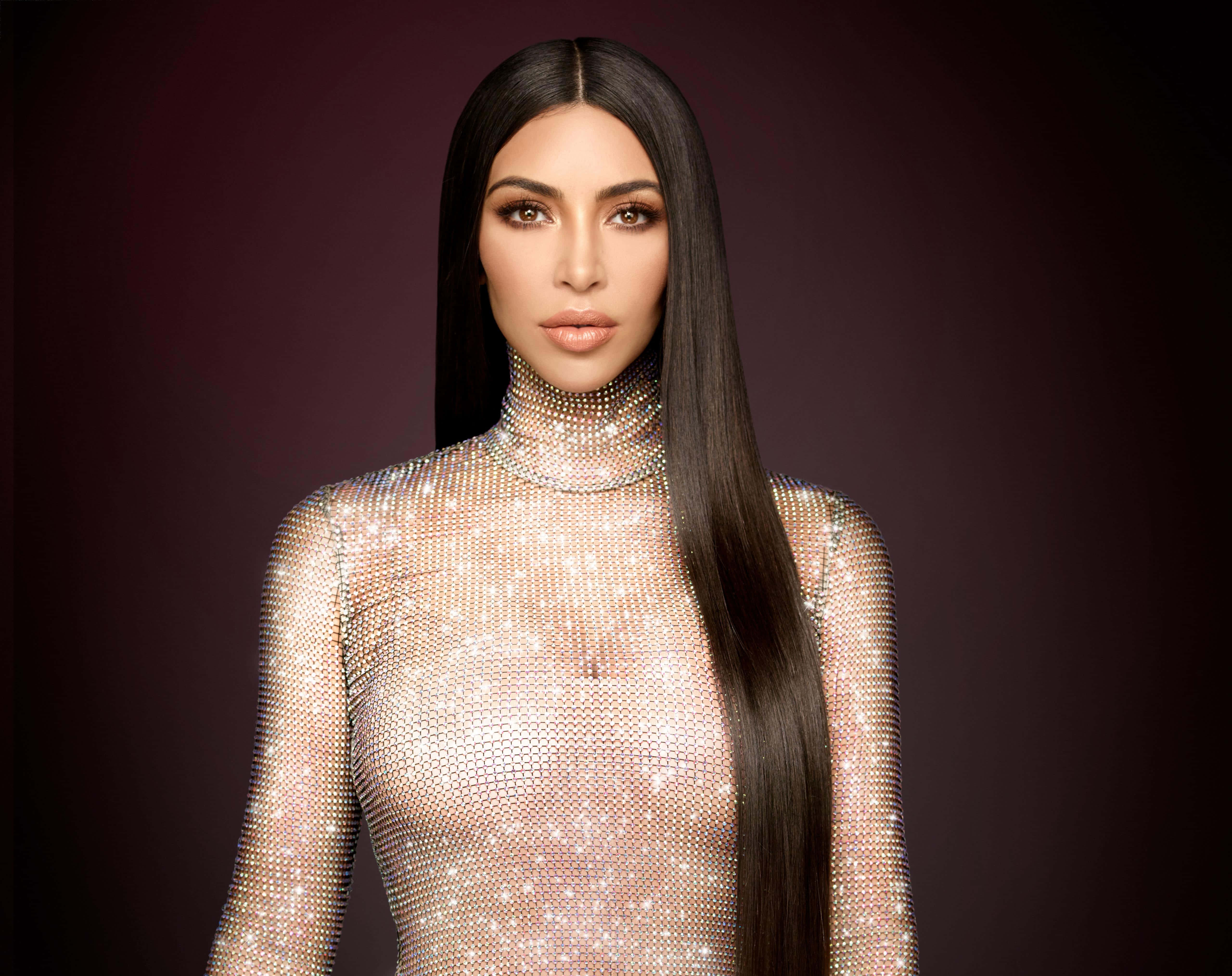 Kim Kardashian 桌布 壁紙 — 一位長黑髮的迷人女性，在深酒紅色背景前穿著閃亮的銀色連體衣。