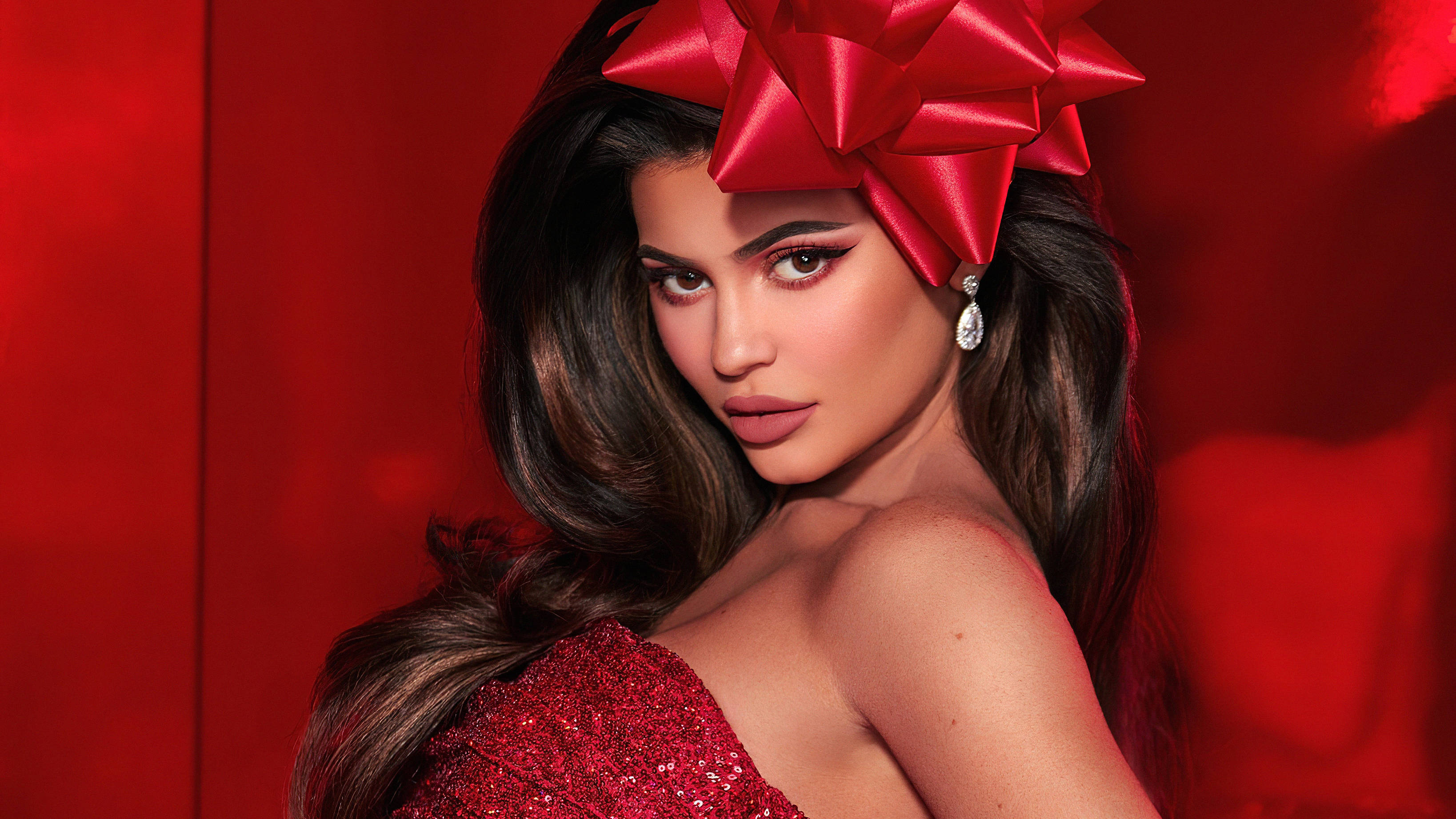 Kylie 桌布 壁紙 — 一位長黑髮的迷人女性，頭上戴著一個大紅色蝴蝶結，穿著閃亮的紅色連衣裙，背景是鮮豔的紅色。