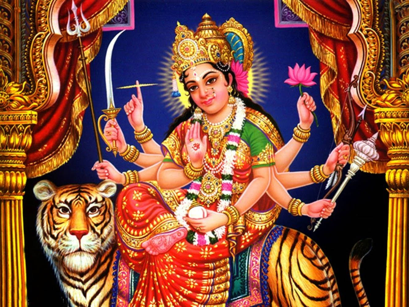 Vaishno Devi 桌布 壁紙 — 一位閃耀的印度教女神，身披金紅裝飾，坐在老虎上，手持神聖武器和蓮花，背景為深藍色。