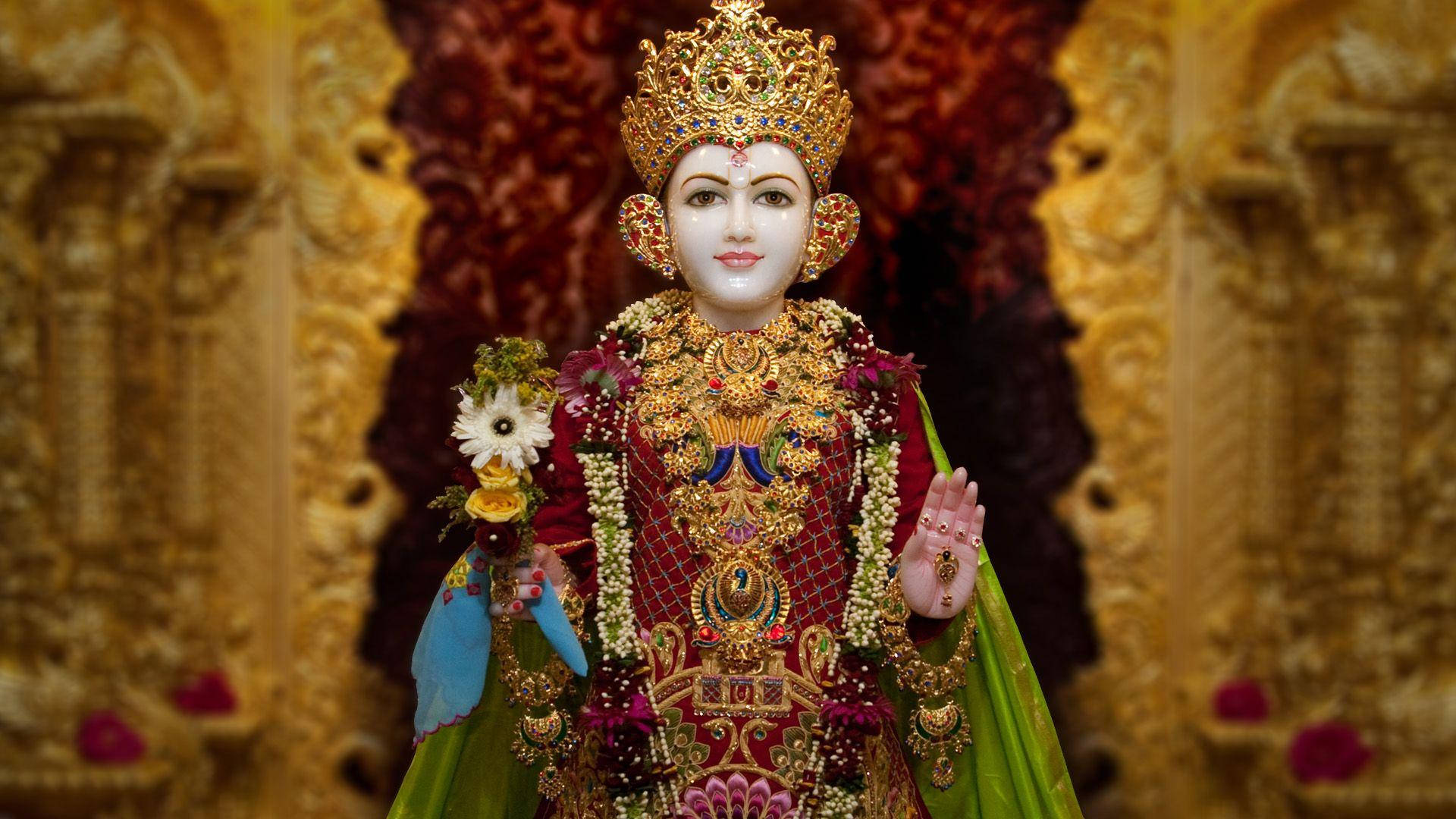 Swaminarayan 桌布 壁紙 — 一位閃耀的神祇，身著精緻的金飾和紅色服飾，站在裝飾華麗的金色拱門前，伴隨著花卉供品。