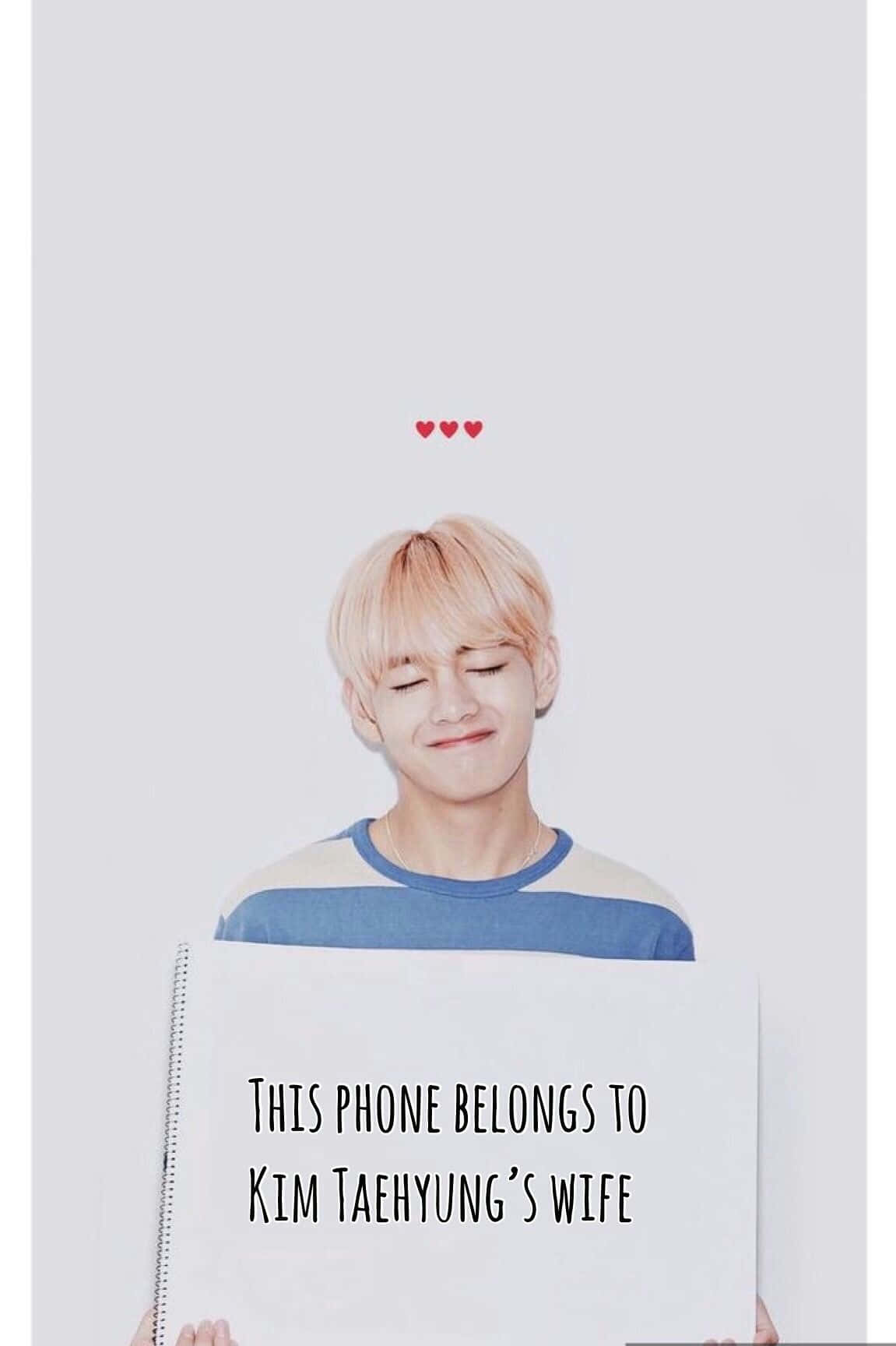 Bts Funny 桌布 壁紙 — 一位閉著眼睛、帶著溫柔微笑的年輕男子，在三顆漂浮的紅心下方舉著一塊牌子，上面寫著「這部電話屬於金泰亨的妻子」。