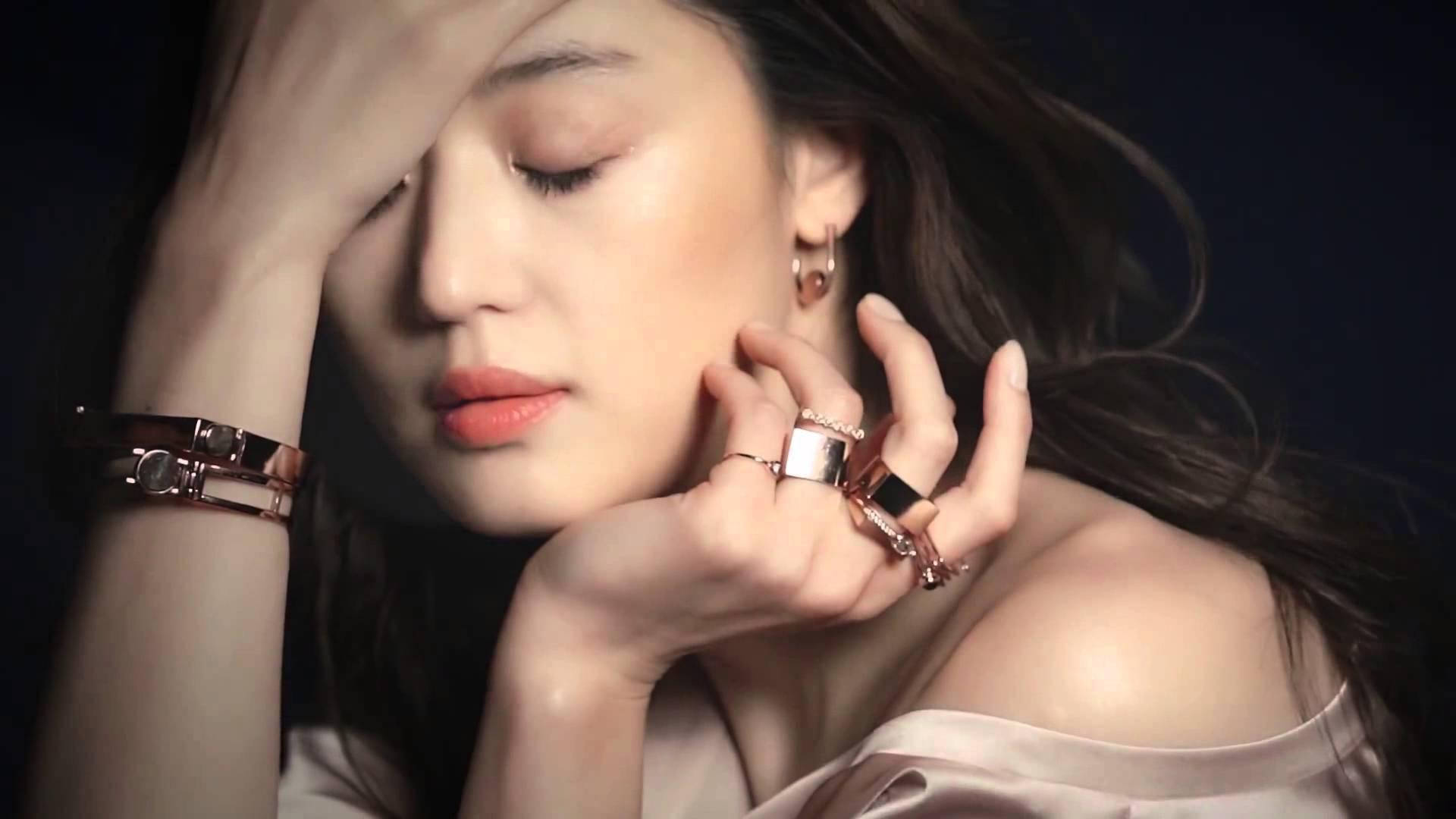 Jun Ji Hyun 桌布 壁紙 — 一位閉著眼睛、秀髮飄逸的女性，在深色背景下輕輕觸摸她戴著玫瑰金首飾的臉龐。