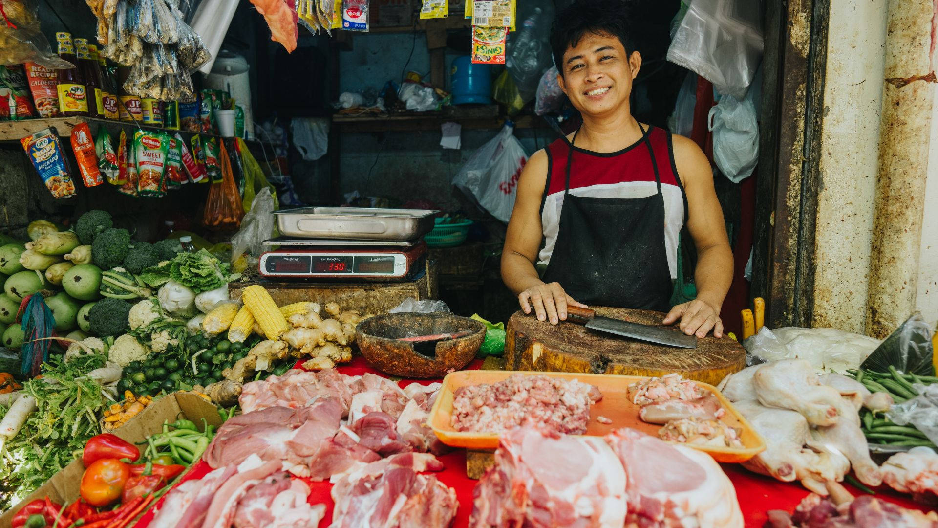 smiling 桌布 壁紙 — 一位開心的屠夫在一個充滿新鮮肉類、蔬菜和色彩繽紛包裝商品的熱鬧攤位後面微笑。