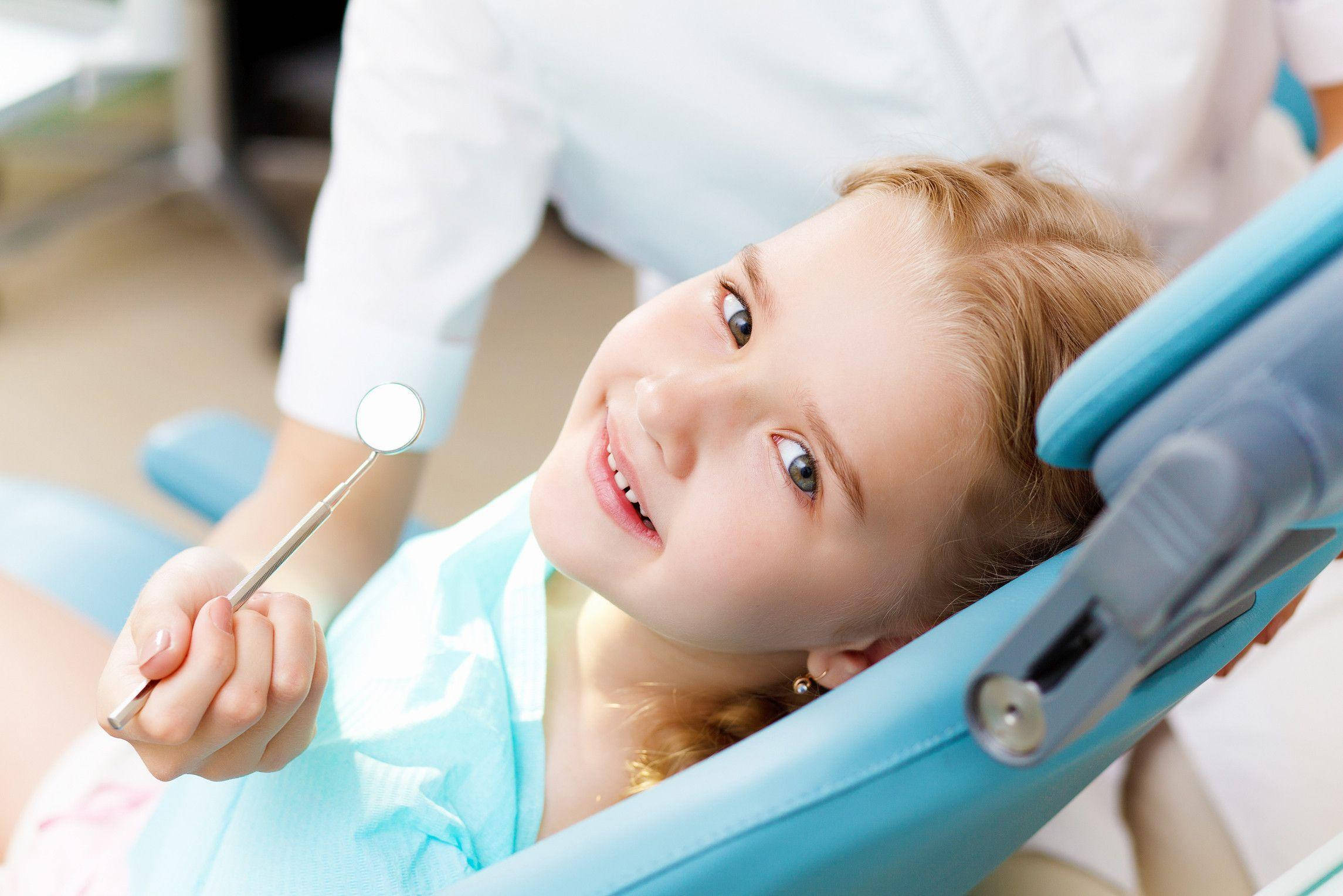 Dentistry 桌布 壁紙 — 一位開心的年輕女孩坐在藍色牙科椅上，手持牙科鏡，露出燦爛的笑容。
