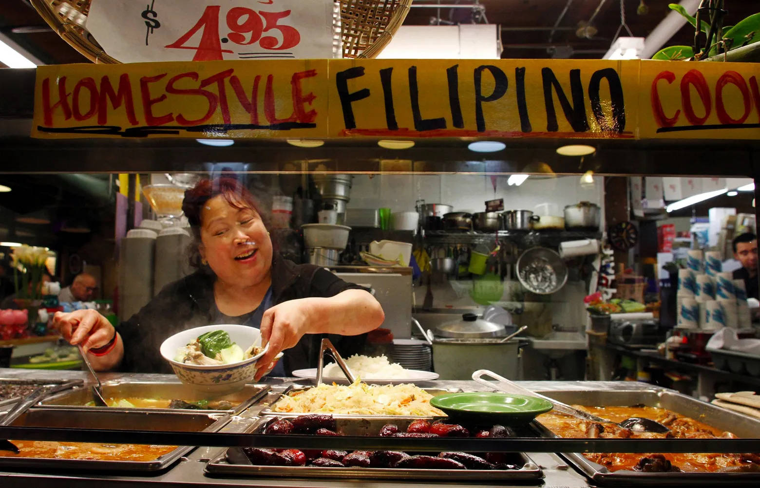 Pike Place Market 桌布 壁紙 — 一位開心的攤販在一個熙熙攘攘、招牌色彩繽紛的美食攤位提供熱氣騰騰的菲律賓菜餚。