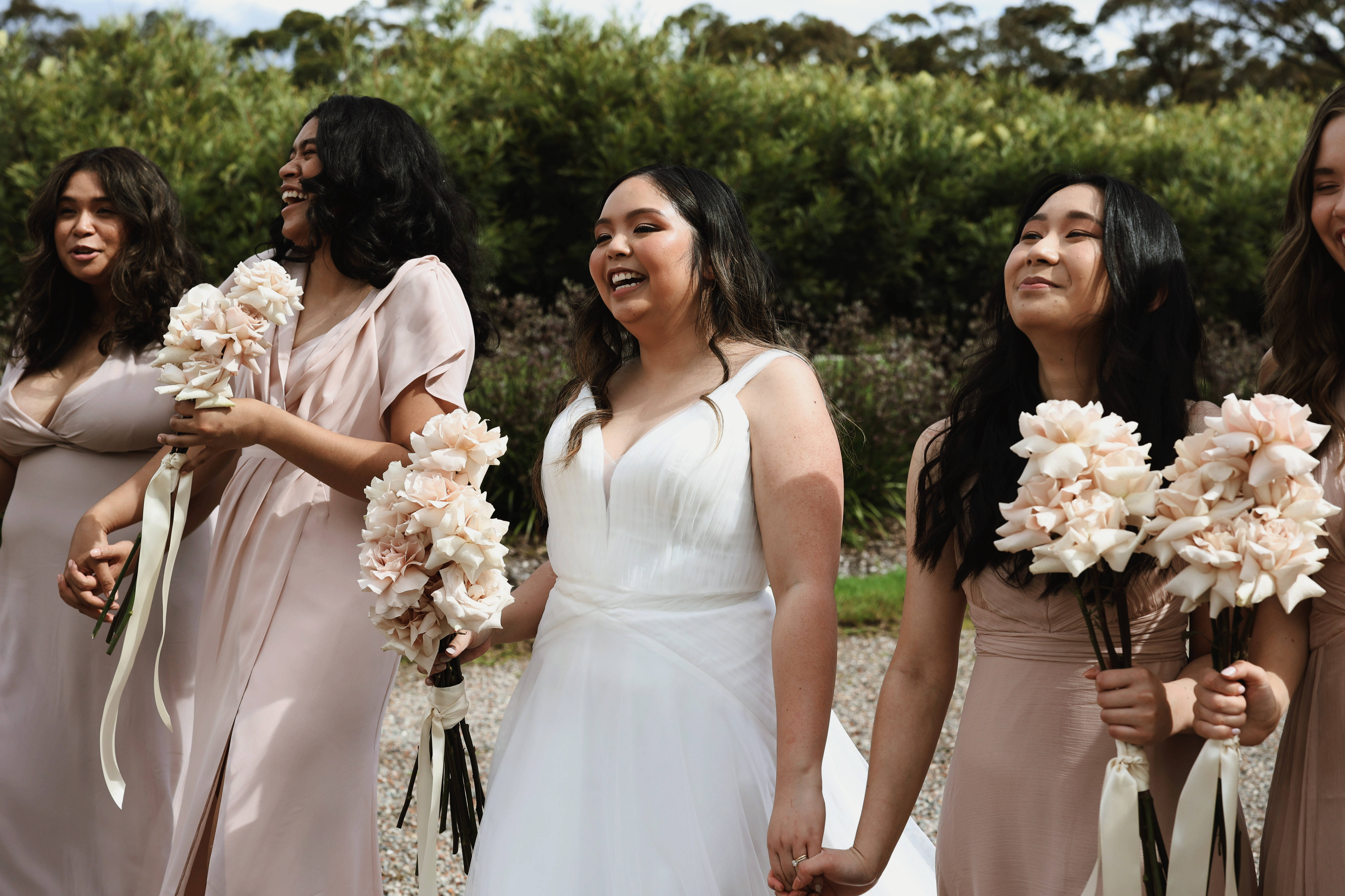 Bridesmaids 桌布 壁紙 — 一位開心的新娘和她的伴娘們穿著淡粉紅色的禮服，在戶外拿著柔和色調的花束一起大笑。