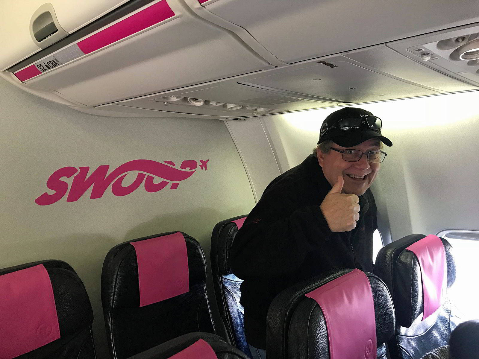 flying 桌布 壁紙 — 一位開心的男士在Swoop飛機艙內，坐在粉紅色和黑色的座位上，豎起大拇指。