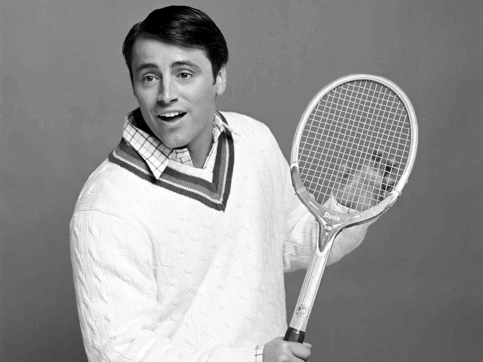 Matt Leblanc 桌布 壁紙 — 一位開心的男士穿著纜繩針織毛衣，在純色工作室背景前拿著一個復古網球拍。