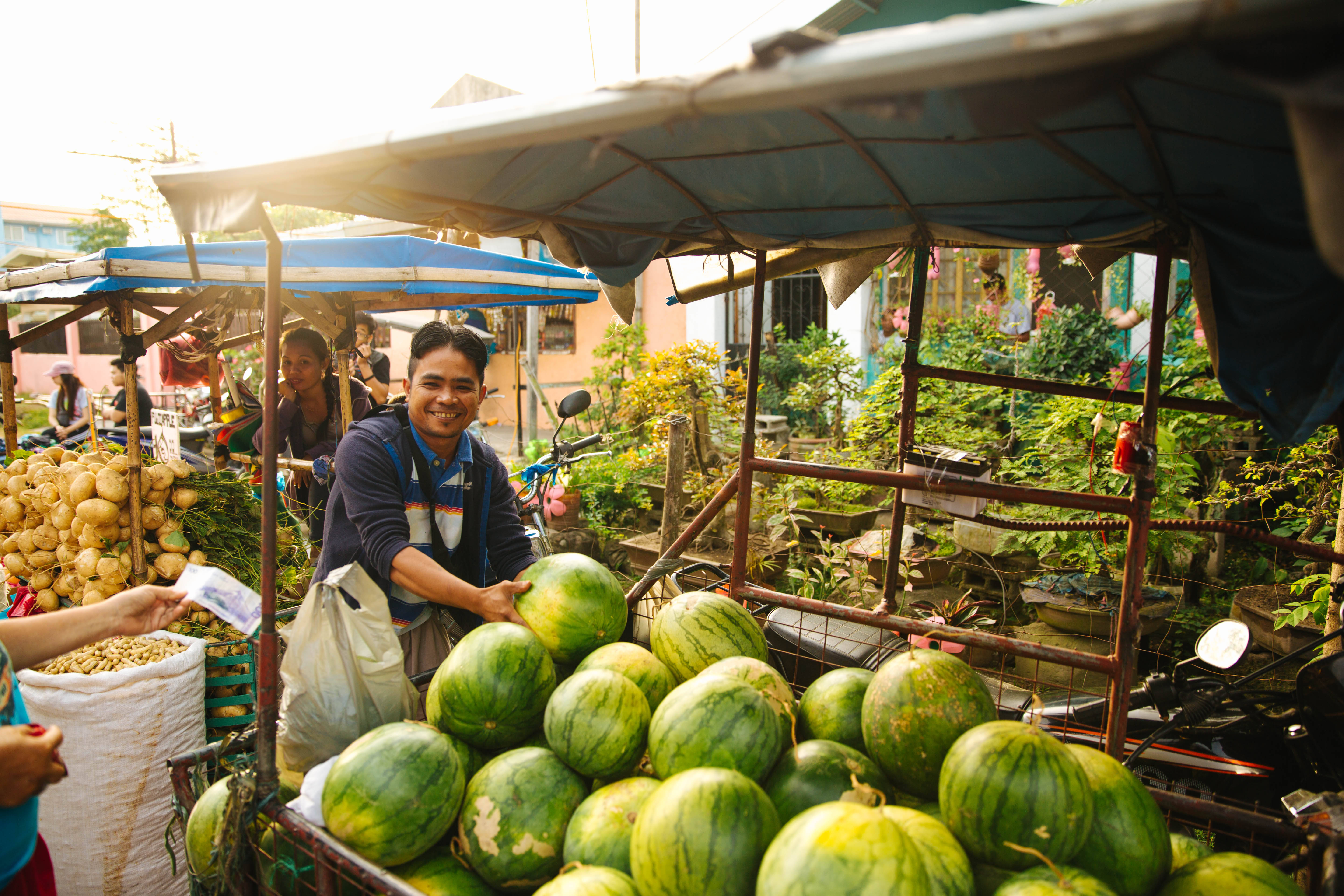 Watermelon 桌布 壁紙 — 一位開心的賣家在天篷下熱鬧的戶外市場攤位賣西瓜時微笑。