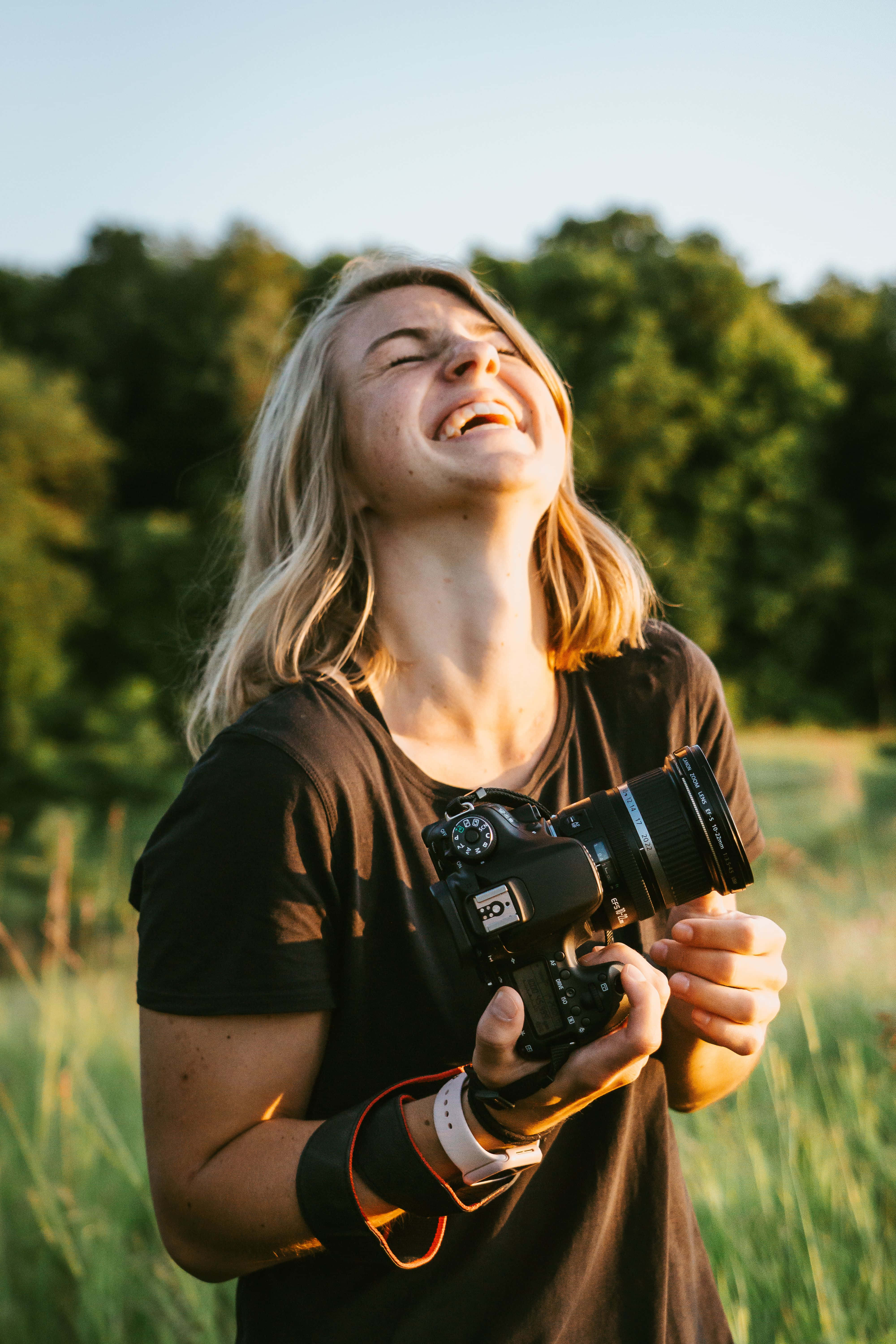 Holding Camera 桌布 壁紙 — 一位開心的金髮女子閉著眼睛笑著，在陽光明媚的草地上拿著單反相機。