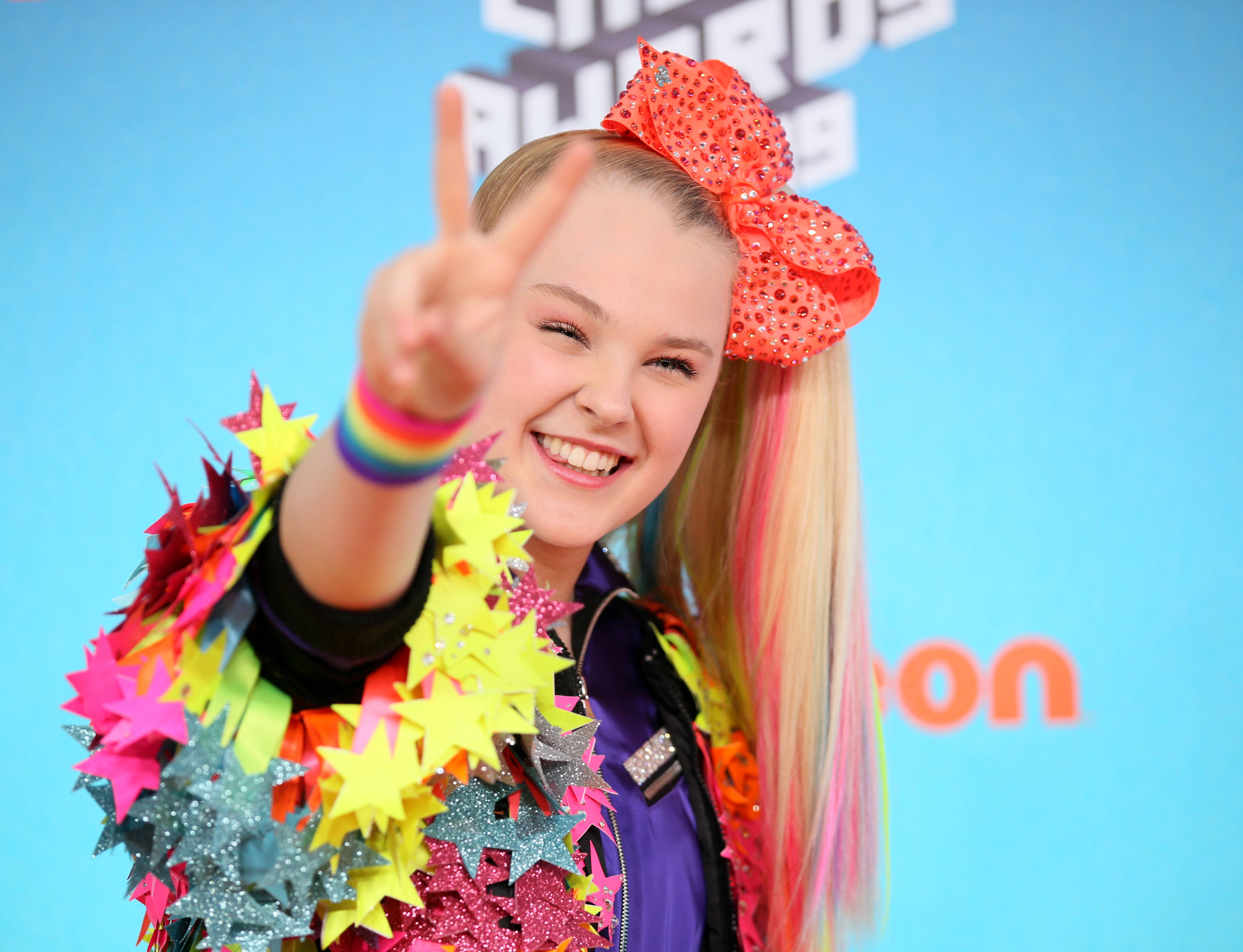 JoJo Siwa 桌布 壁紙 — 一位開朗的小女孩戴著一個巨大的閃亮紅色蝴蝶結，對著鏡頭露出燦爛的笑容並比出和平手勢。