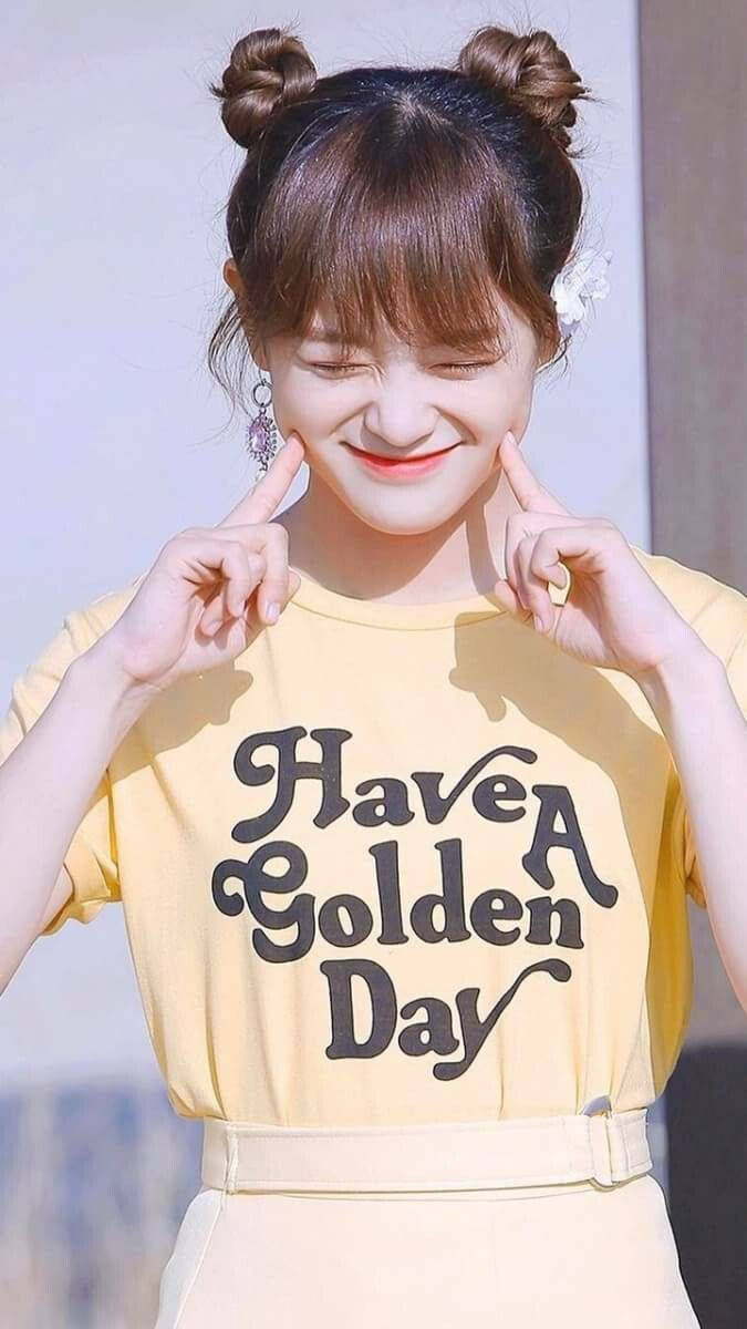 桌布 壁紙 — 一位開朗的年輕女性，梳著雙丸子頭，身穿黃色「Have A Golden Day」T恤，微笑著調皮地觸摸臉頰。