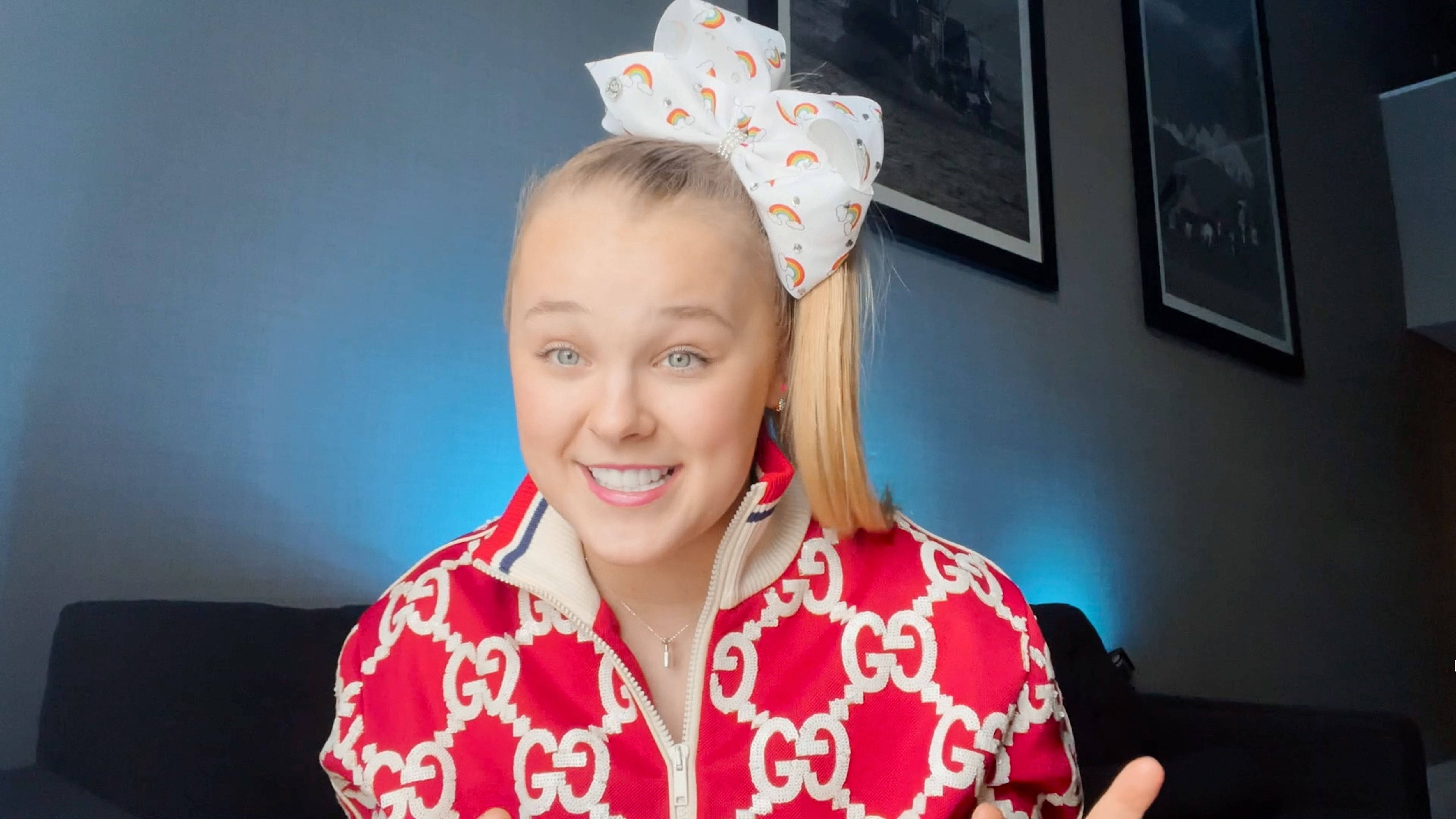 JoJo Siwa 桌布 壁紙 — 一位開朗的年輕女性，金髮紮成馬尾，戴著一個大彩虹蝴蝶結，身穿紅色GG圖案外套，在室內對著鏡頭微笑。