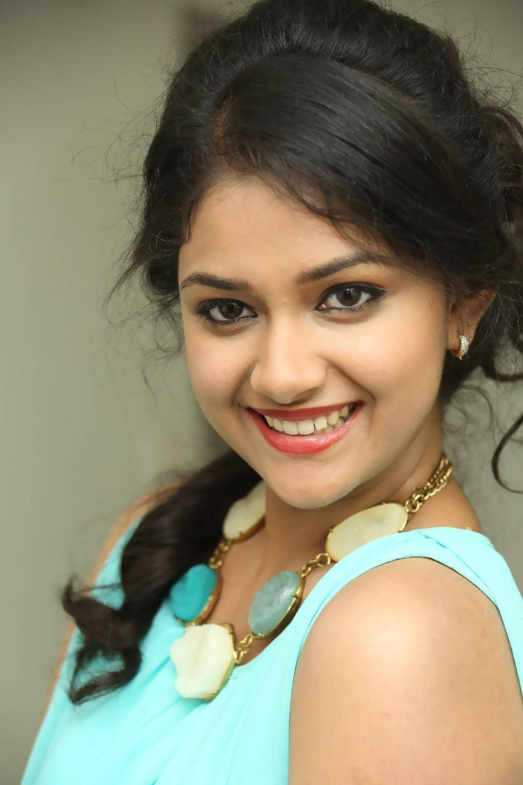Keerthi Suresh HD 桌布 壁紙 — 一位開朗的年輕黑髮女性，穿著藍綠色上衣和一條個性項鍊，露出燦爛的笑容。