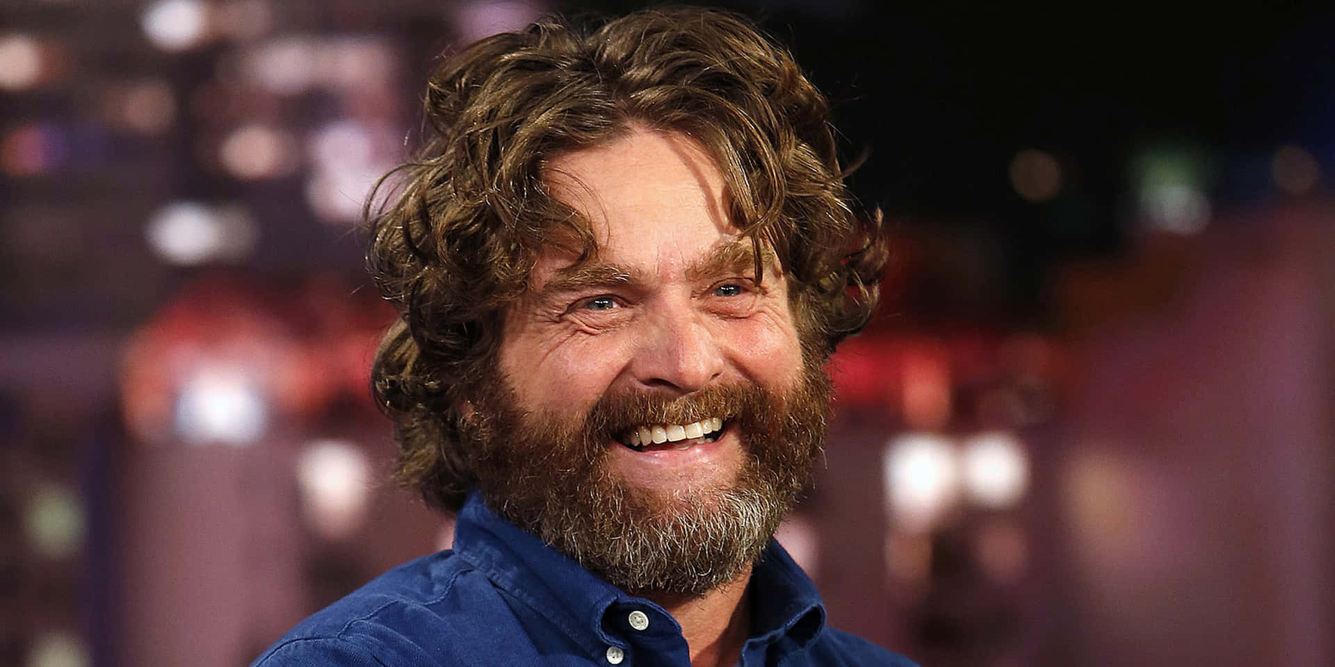 Zach Galifianakis 桌布 壁紙 — 一位開朗的男子，留著棕色捲髮和濃密鬍鬚，身穿藍色襯衫，在模糊的城市夜景背景下溫暖地微笑。