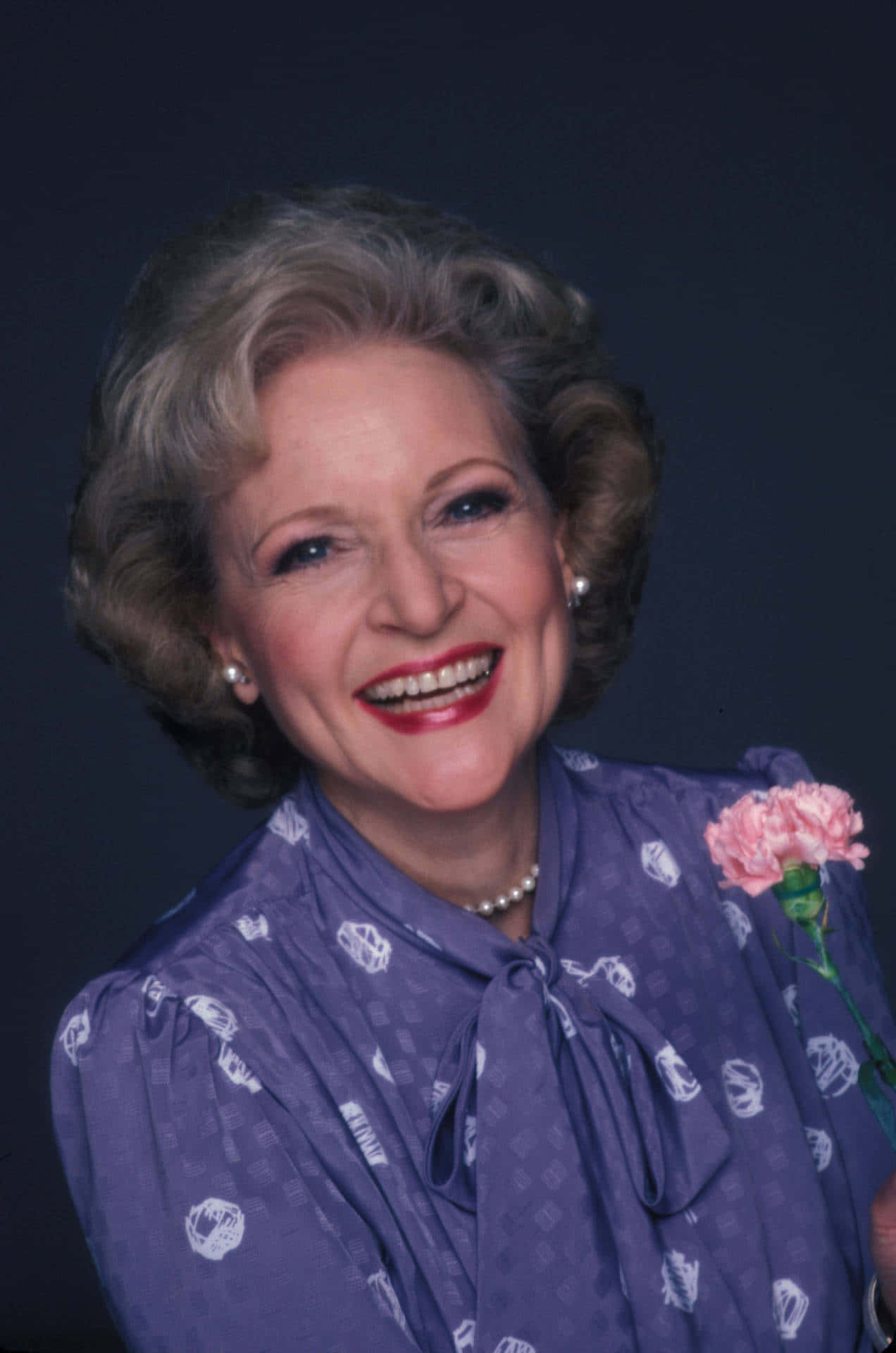 Betty White 桌布 壁紙 — 一位開朗的白髮老婦人，在深色背景下手持粉紅色康乃馨，露出燦爛的笑容。