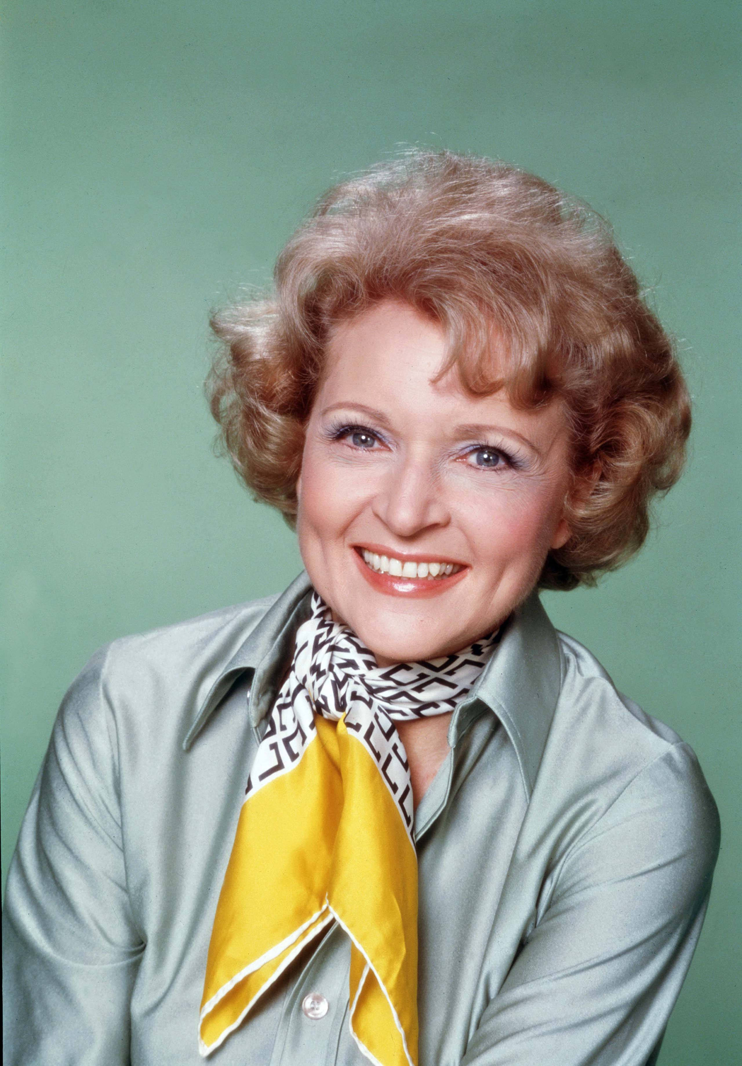 Betty White 桌布 壁紙 — 一位開朗的金色捲髮女性，身穿銀色襯衫和黃色圖案圍巾，在綠色背景前溫暖地微笑。