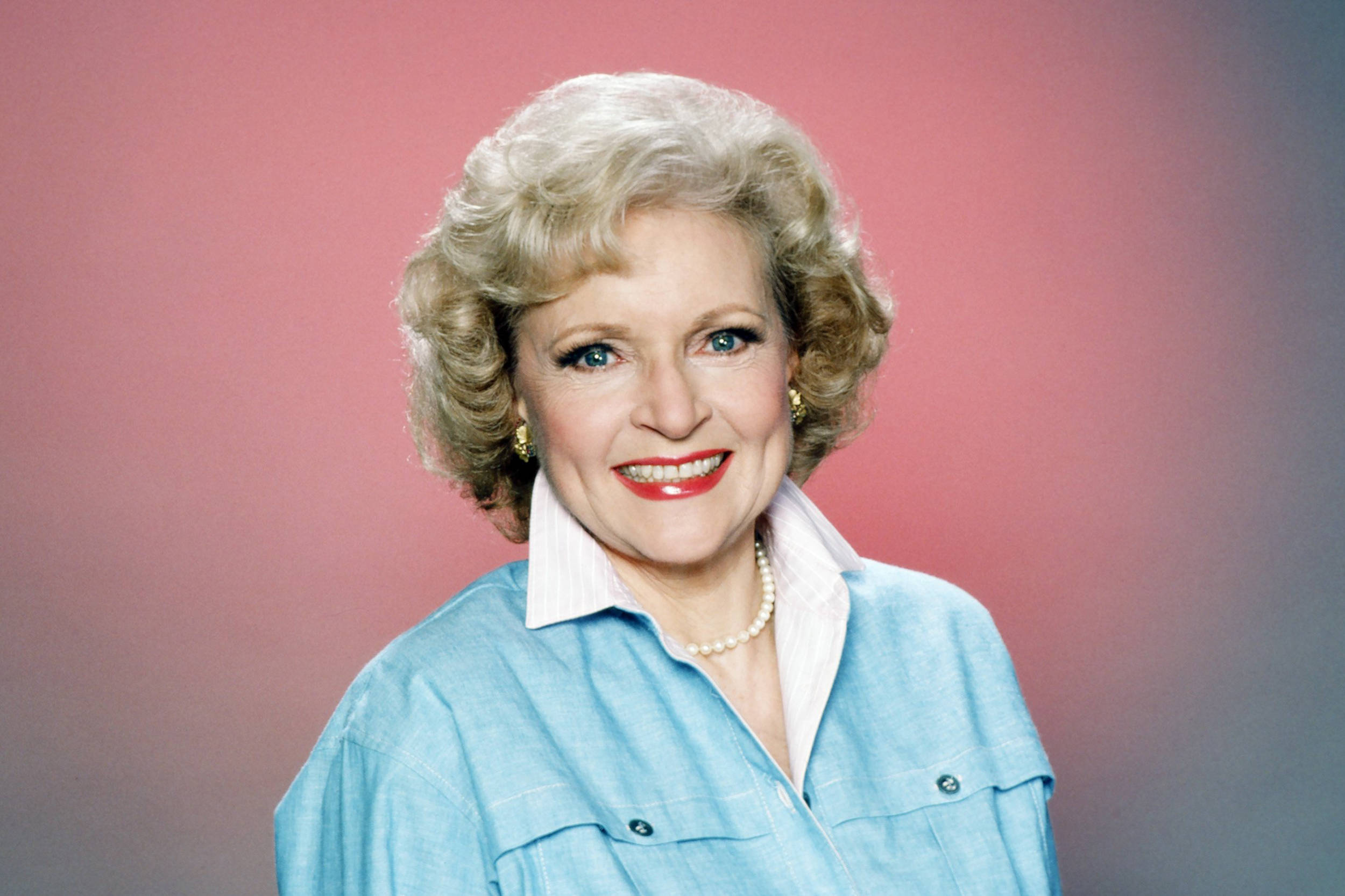 Betty White 桌布 壁紙 — 一位開朗的金色捲髮紅唇女性在柔和的粉紅色漸變背景前溫暖地微笑。