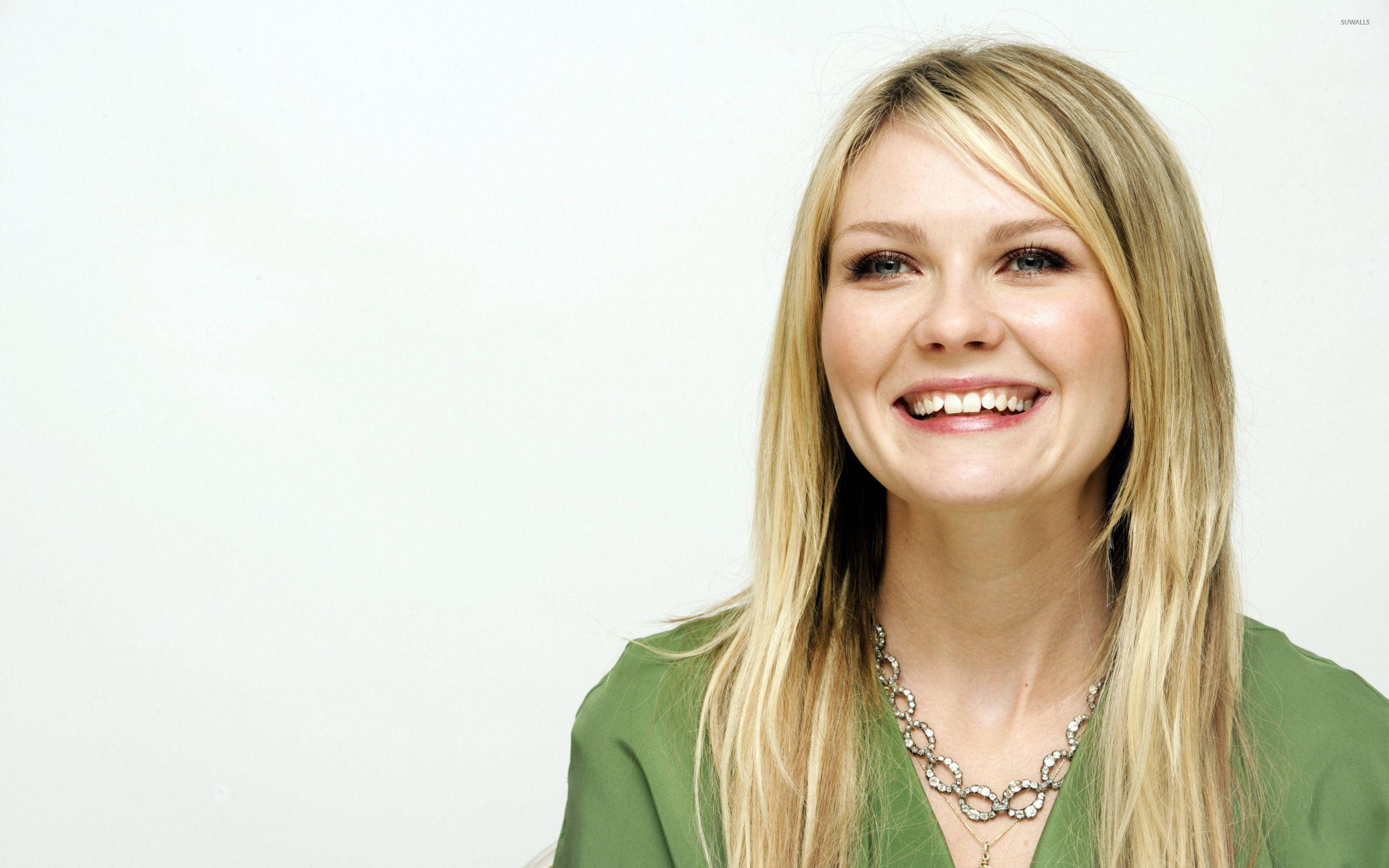 Kirsten Dunst 桌布 壁紙 — 一位開朗的金髮女性，面帶燦爛笑容，穿著綠色上衣和銀色項鍊，背景為純白色。