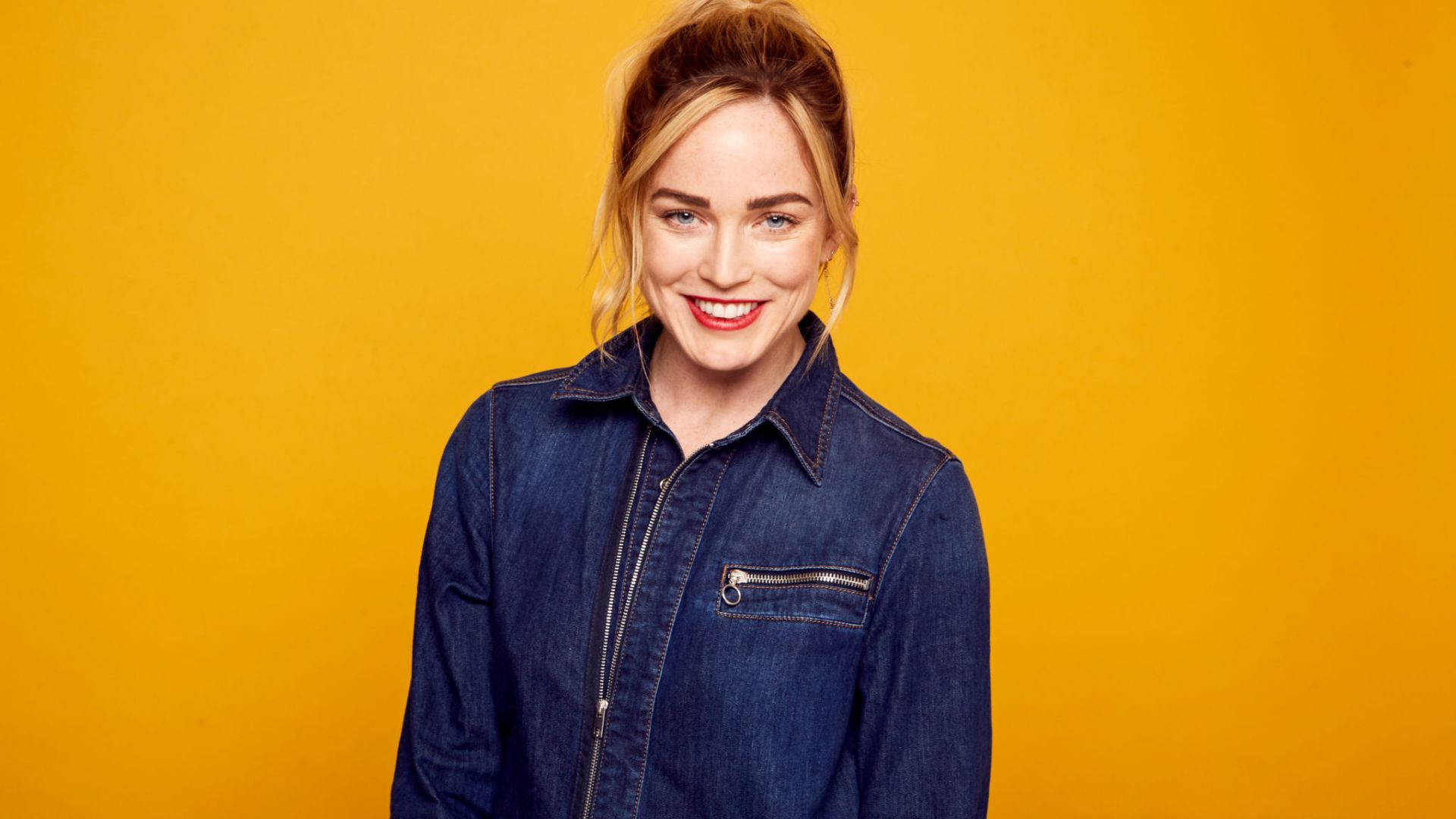 一位微笑的女性在黃色背景下穿著牛仔外套。 — Caity Lotz 動圖 壁紙