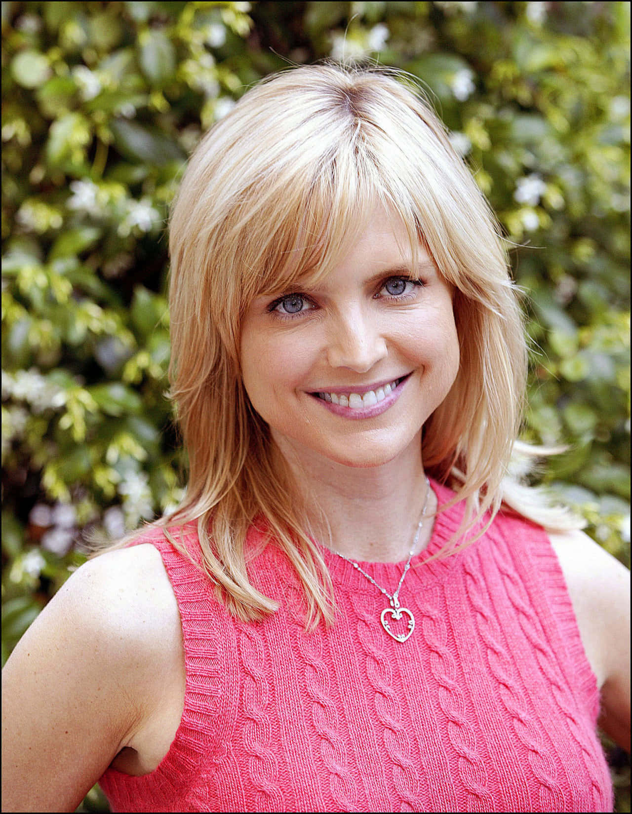 Courtney Thorne Smith 桌布 壁紙 — 一位開朗的金髮女性穿著粉紅色麻花針織上衣，在茂密的綠色樹葉背景下溫暖地微笑。