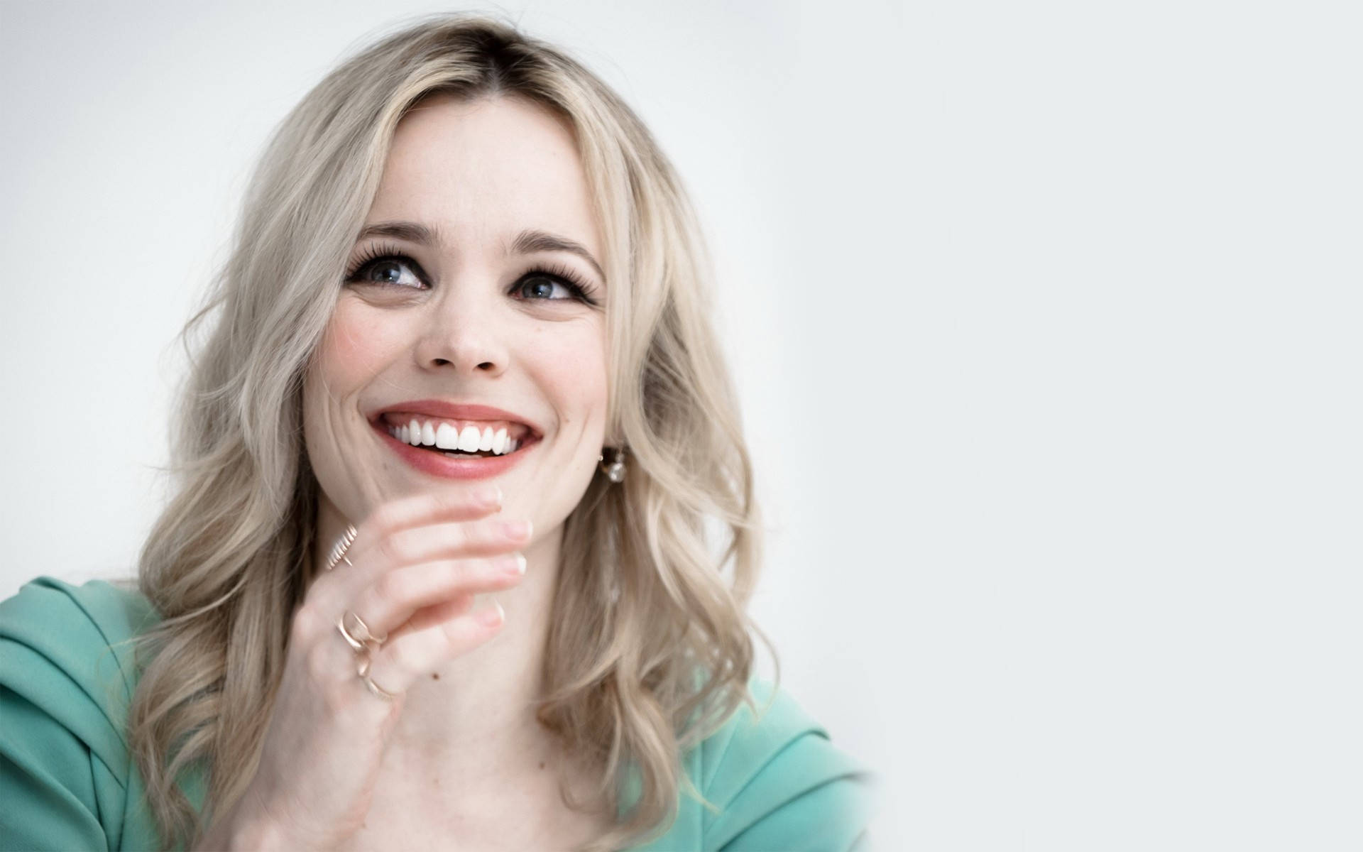 Rachel Mcadams 桌布 壁紙 — 一位開朗的金髮女性穿著青綠色上衣，在純白背景前微笑著輕輕觸碰下巴。