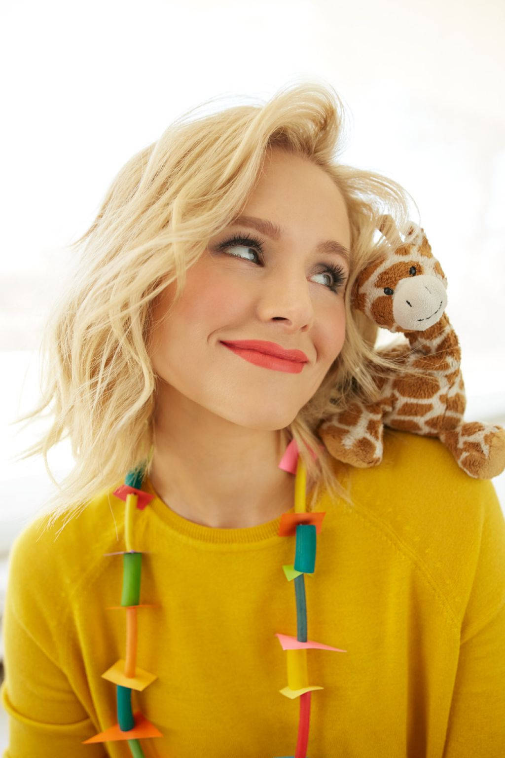 kristen bell 桌布 壁紙 — 一位開朗的金髮女性穿著黃色毛衣，微笑著抬頭抱著長頸鹿毛絨玩具。