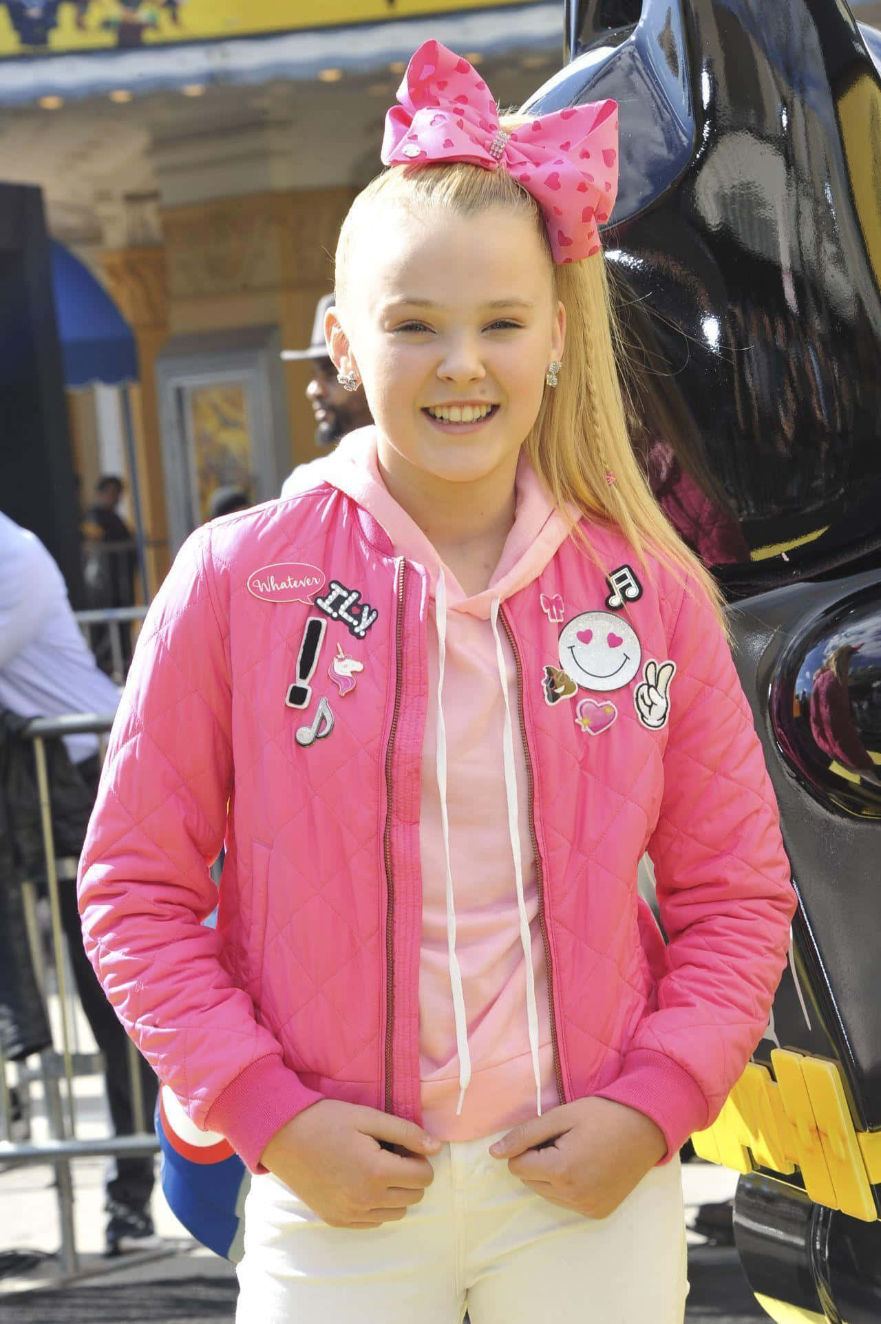 JoJo Siwa 桌布 壁紙 — 一位開朗的金髮年輕女孩，戴著巨大的粉紅色蝴蝶結，穿著一件飾有彩色補丁的鮮豔粉紅色絎縫外套，露出燦爛的笑容。